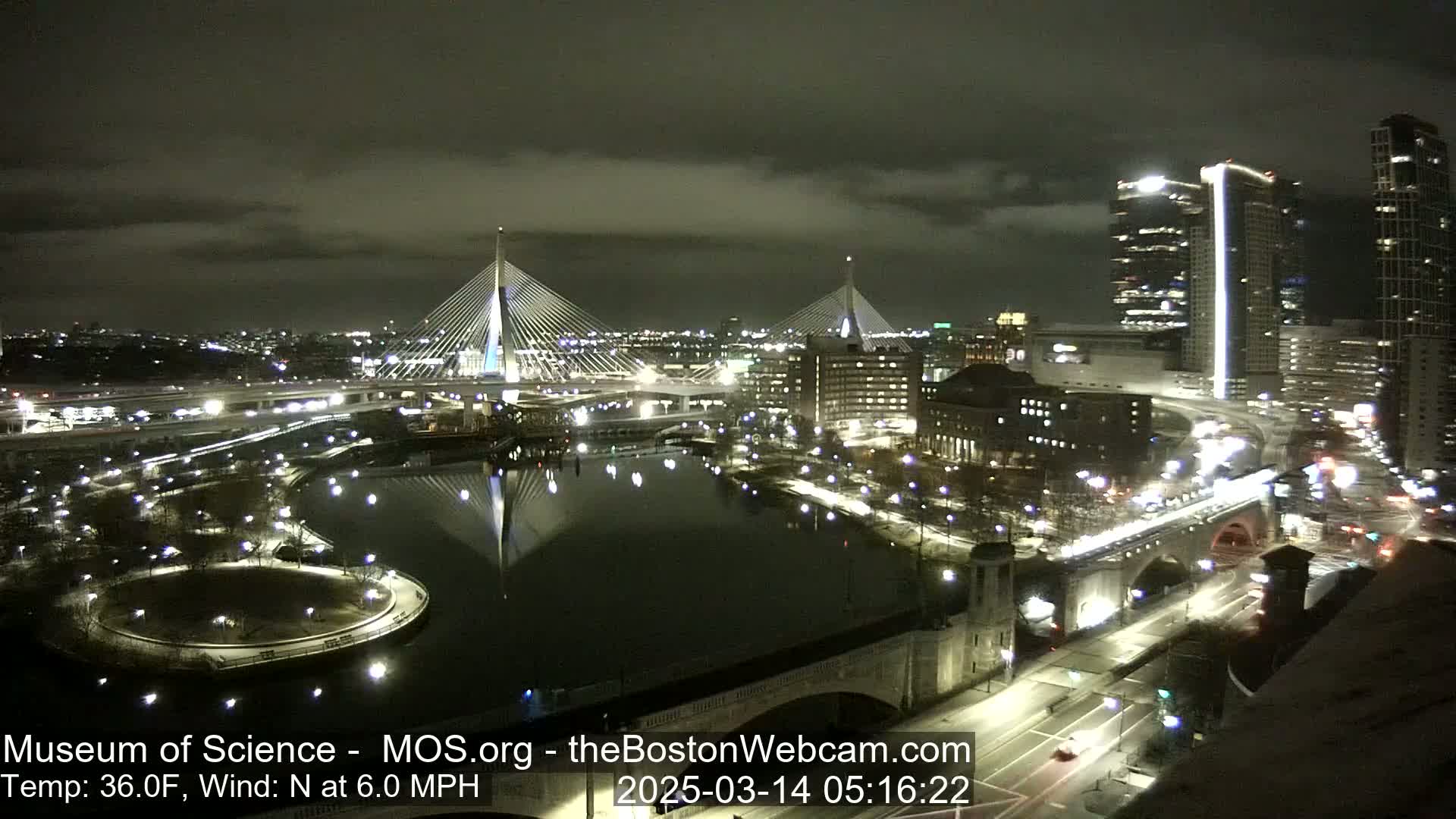 A nighttime cityscape with two cable-stayed bridges reflected in a dark river under a partly cloudy sky.