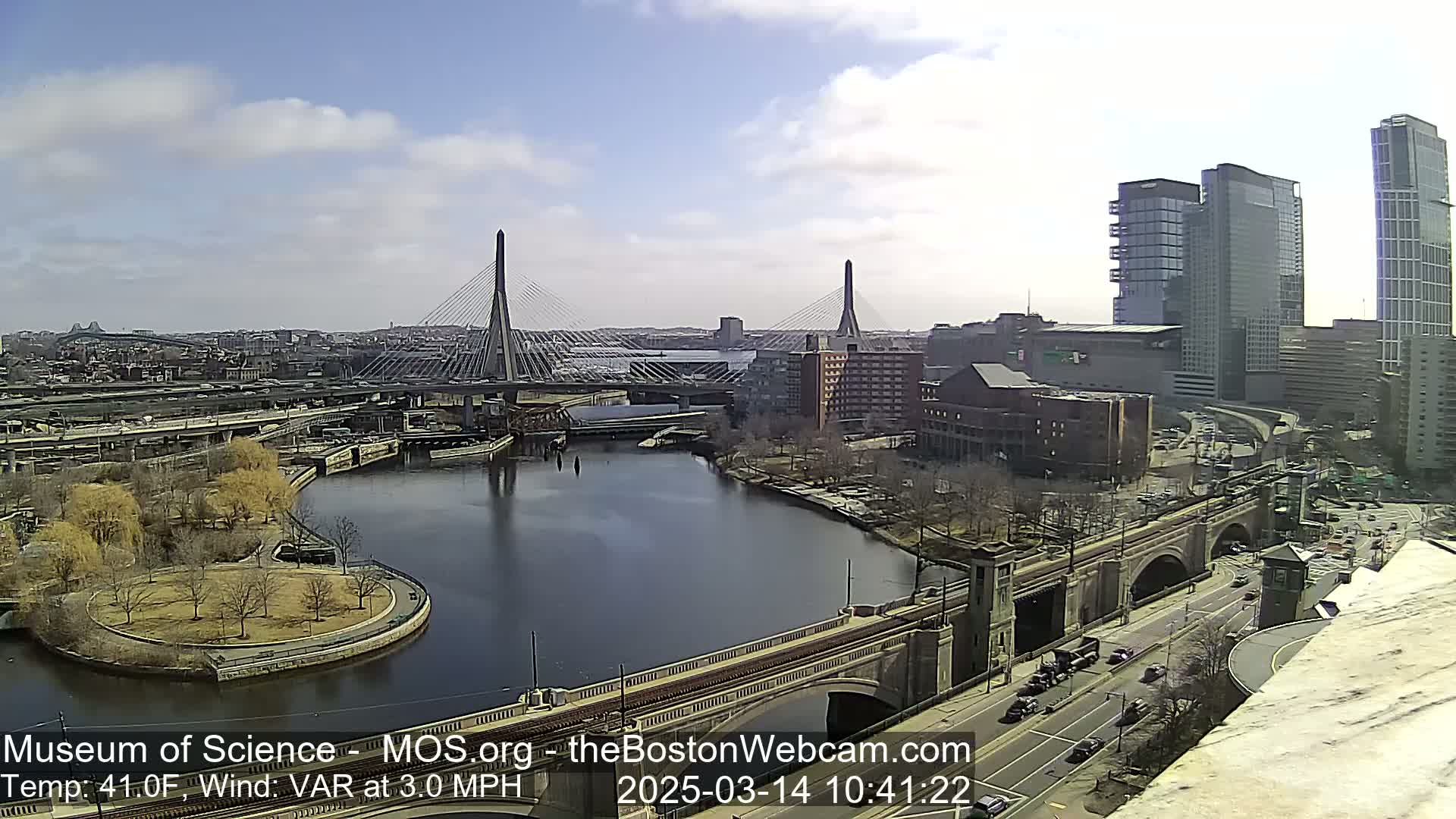 A sunny cityscape with two cable-stayed bridges spanning a river, surrounded by buildings, roadways, and a park.