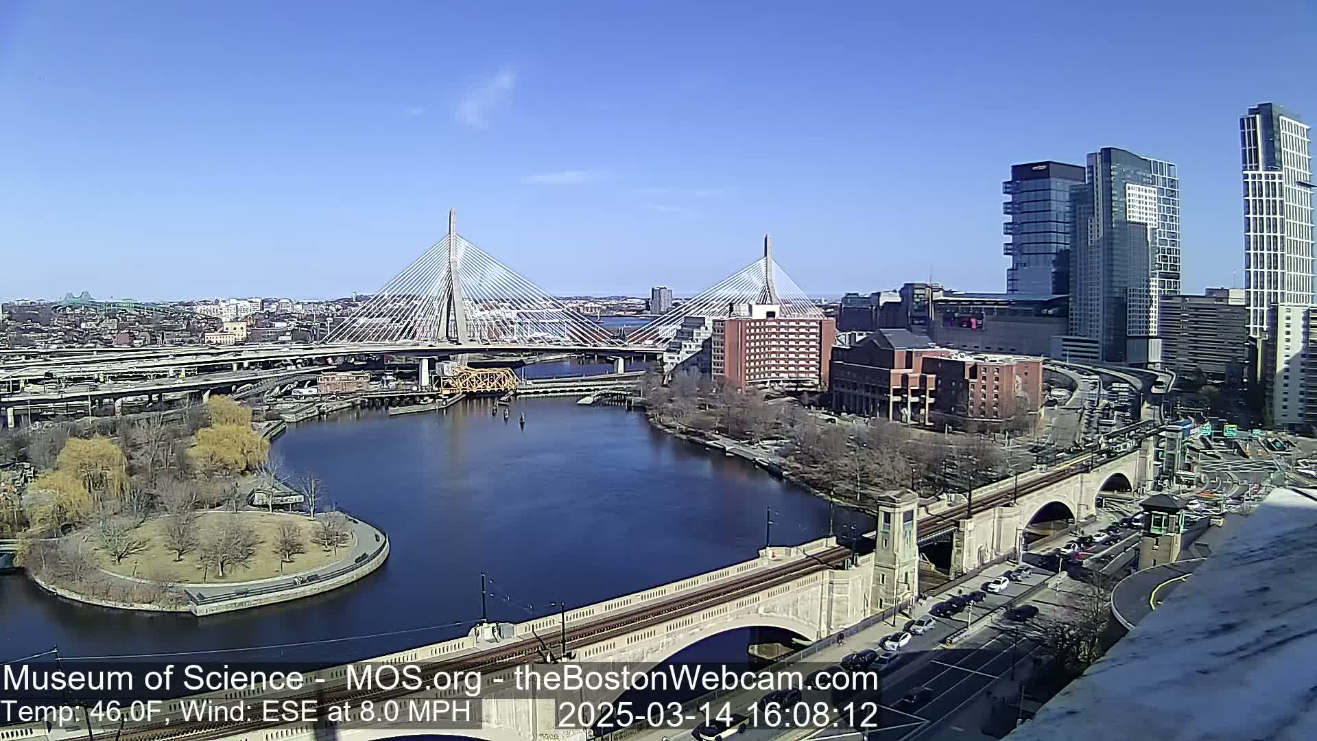 A sunny cityscape shows two cable-stayed bridges spanning a river, with city buildings and roadways visible under a clear blue sky.
