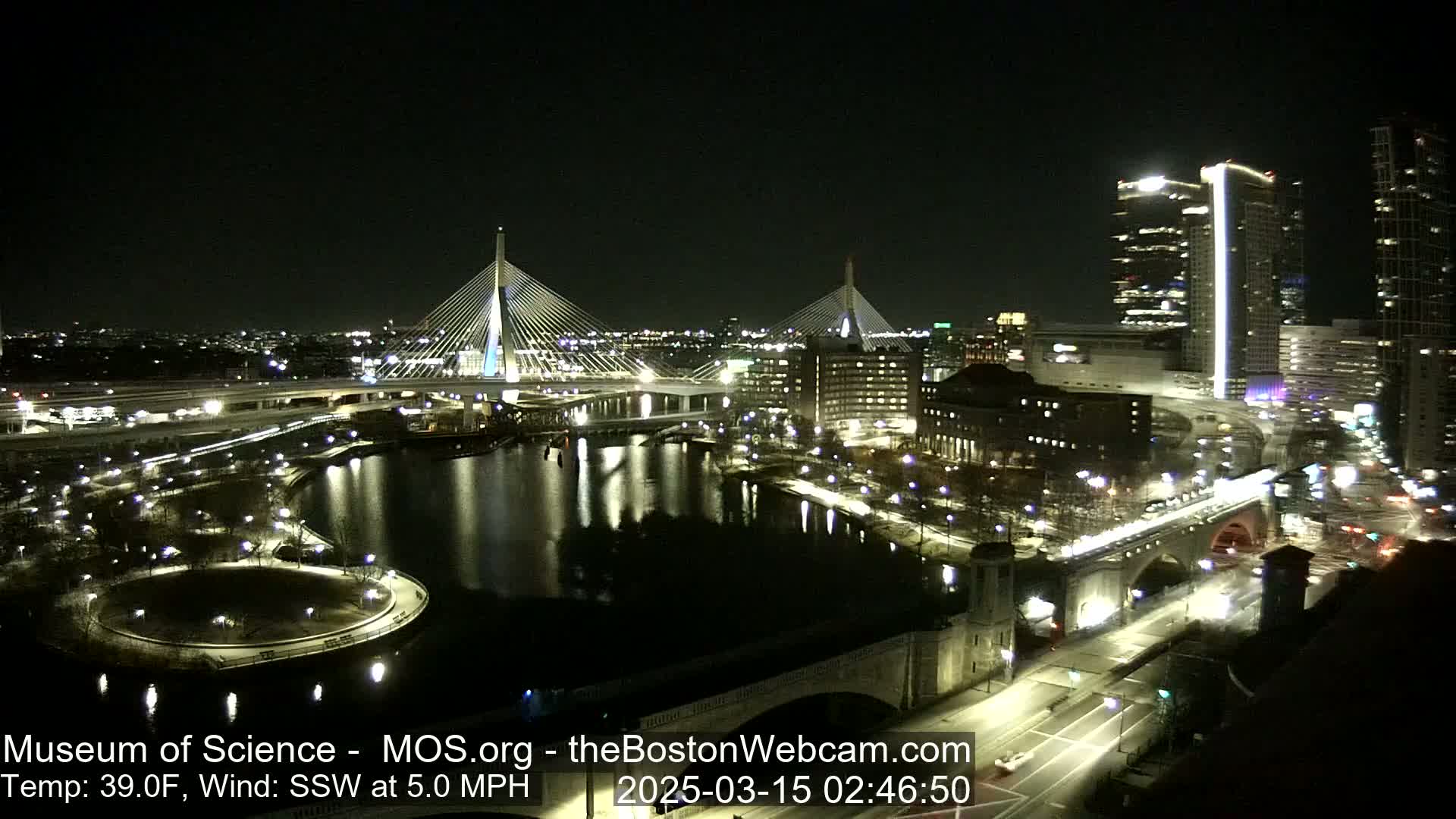 A nighttime view of illuminated bridges and city buildings along a dark river, under a clear sky with a 39°F temperature and a 5.0 mph south-southwesterly wind.
