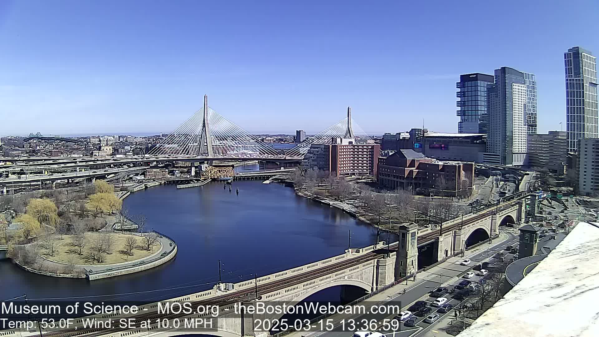 A sunny cityscape shows a river curving around a park, with two cable-stayed bridges and other bridges visible, along with numerous buildings and roadways.