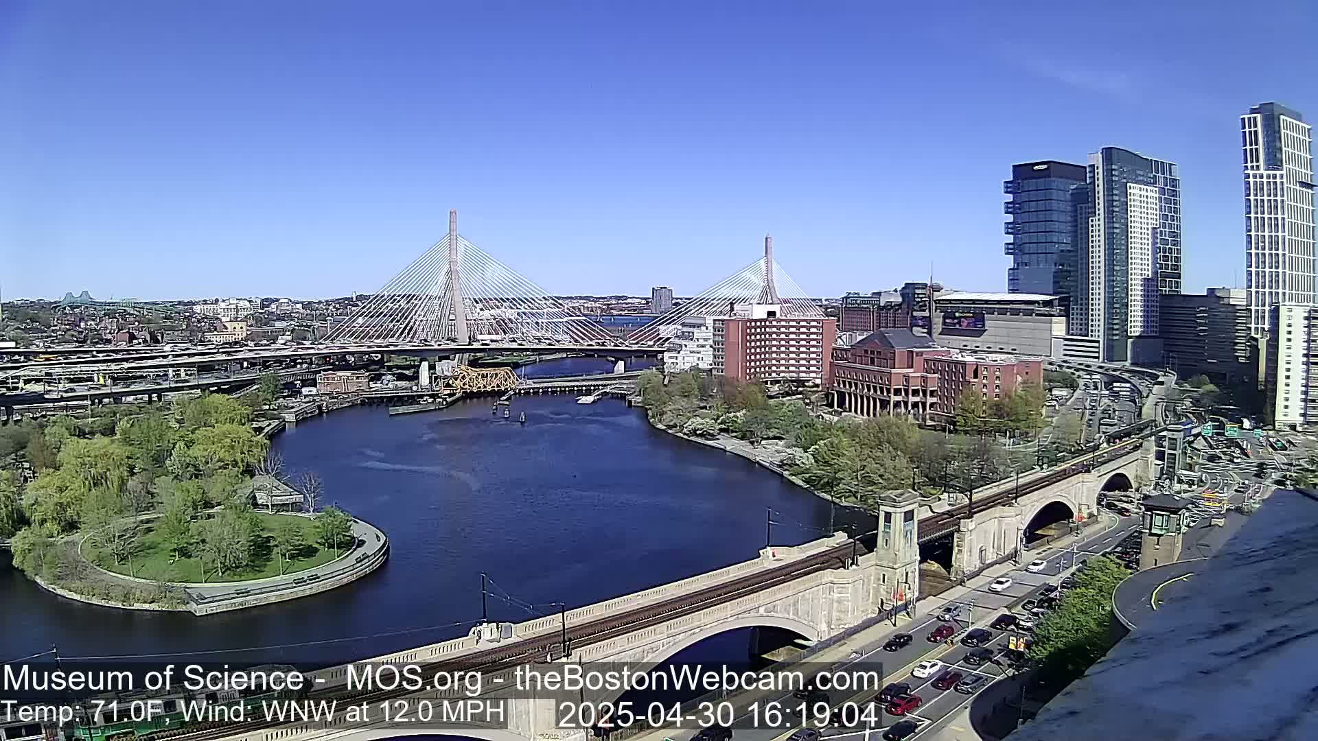 A sunny cityscape featuring two cable-stayed bridges spanning a river, with a  bridge and modern high-rises in the foreground, under a clear blue sky and a strong westerly wind.