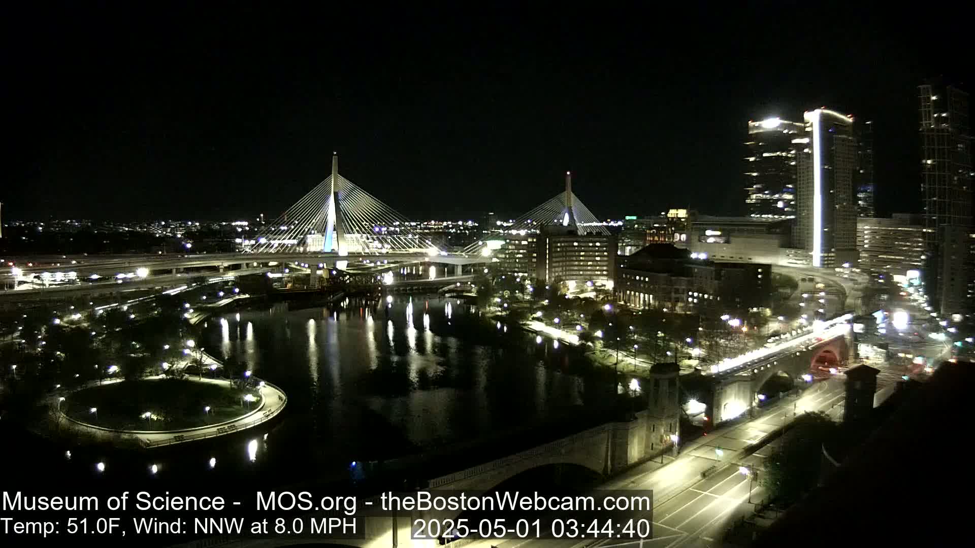 A nighttime cityscape features two cable-stayed bridges spanning a dark river, with city lights reflecting on the water under a clear sky, a cool temperature, and a gentle north-northwest wind.