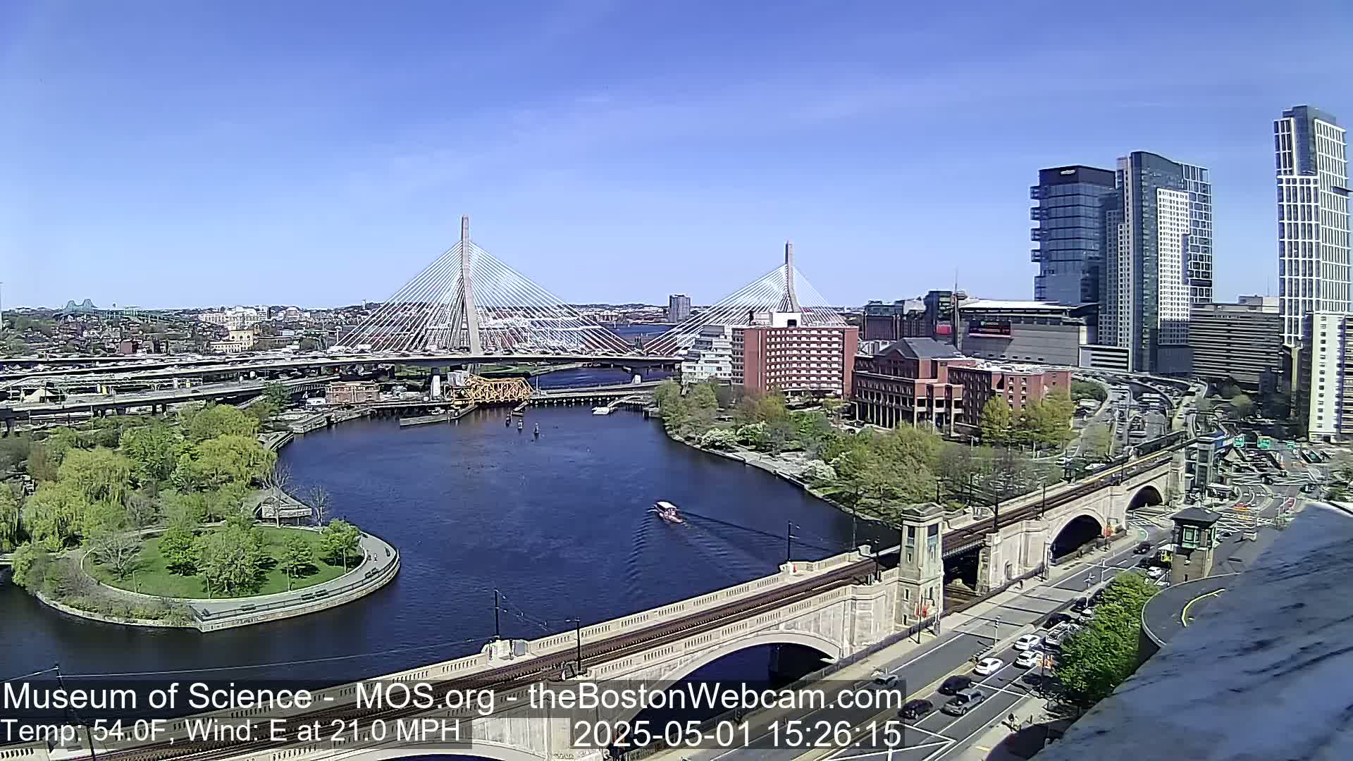 A sunny cityscape shows two cable-stayed bridges spanning a river, with a park on an island in the river, under a clear blue sky and a moderate east wind.
