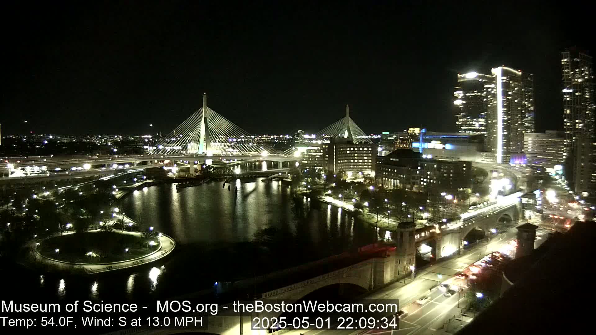 A nighttime cityscape shows two cable-stayed bridges spanning a river, with city lights and traffic visible, under clear skies and a 54°F temperature with a 13 mph south wind.