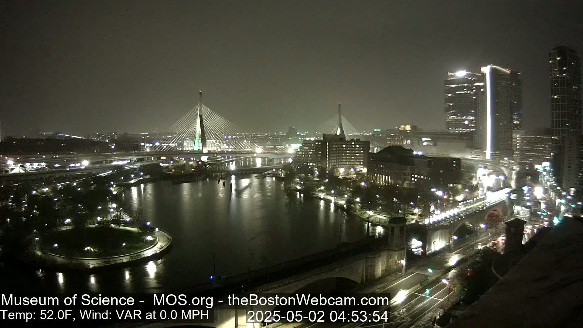 A nighttime view of two cable-stayed bridges and a city skyline across a dark river under a cloudy sky.