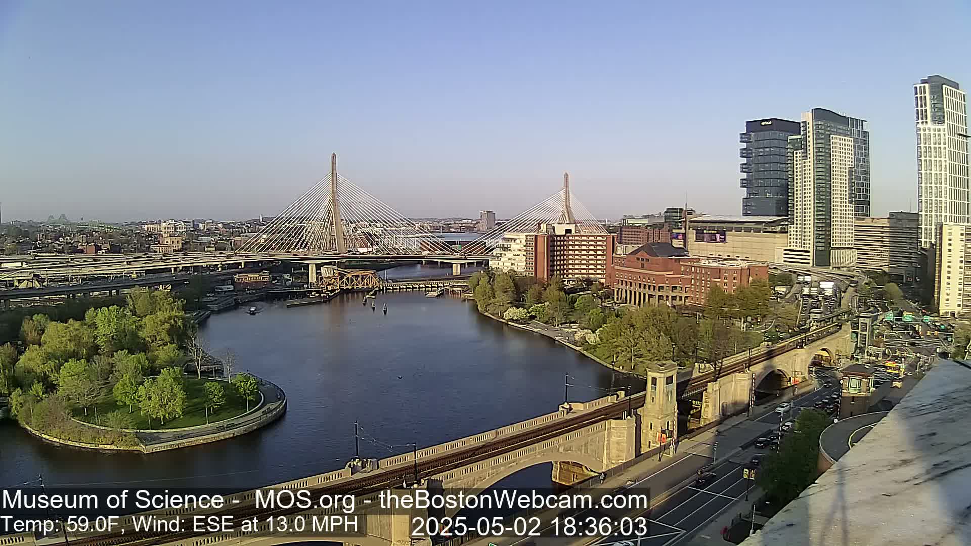 A clear, sunny day reveals a cityscape with two cable-stayed bridges spanning a river, a small island with green trees, and modern high-rise buildings.