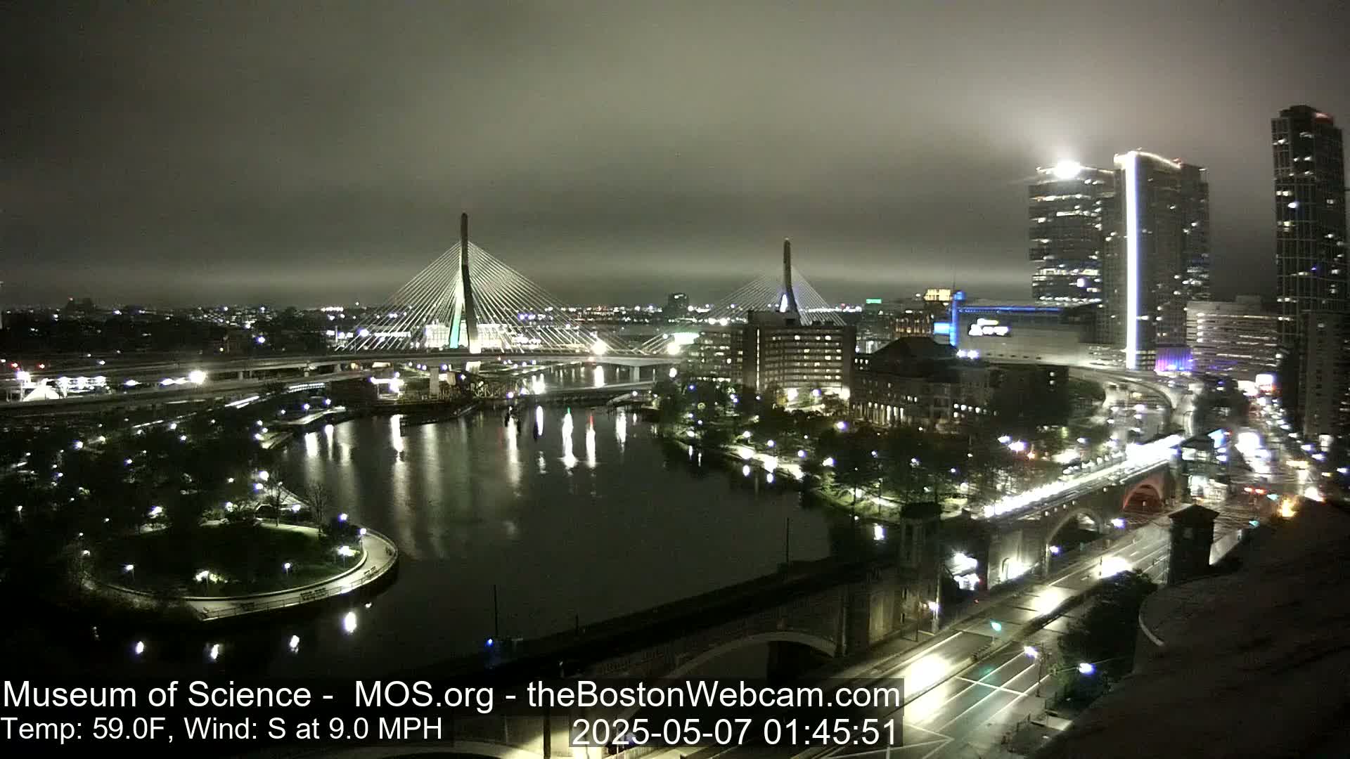 A nighttime cityscape with two cable-stayed bridges illuminated over a dark river, under a foggy sky, with a moderate south wind.
