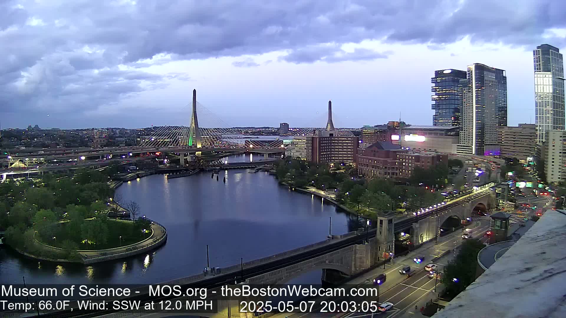 An evening view of a city with two cable-stayed bridges over a river, under a mostly cloudy sky.