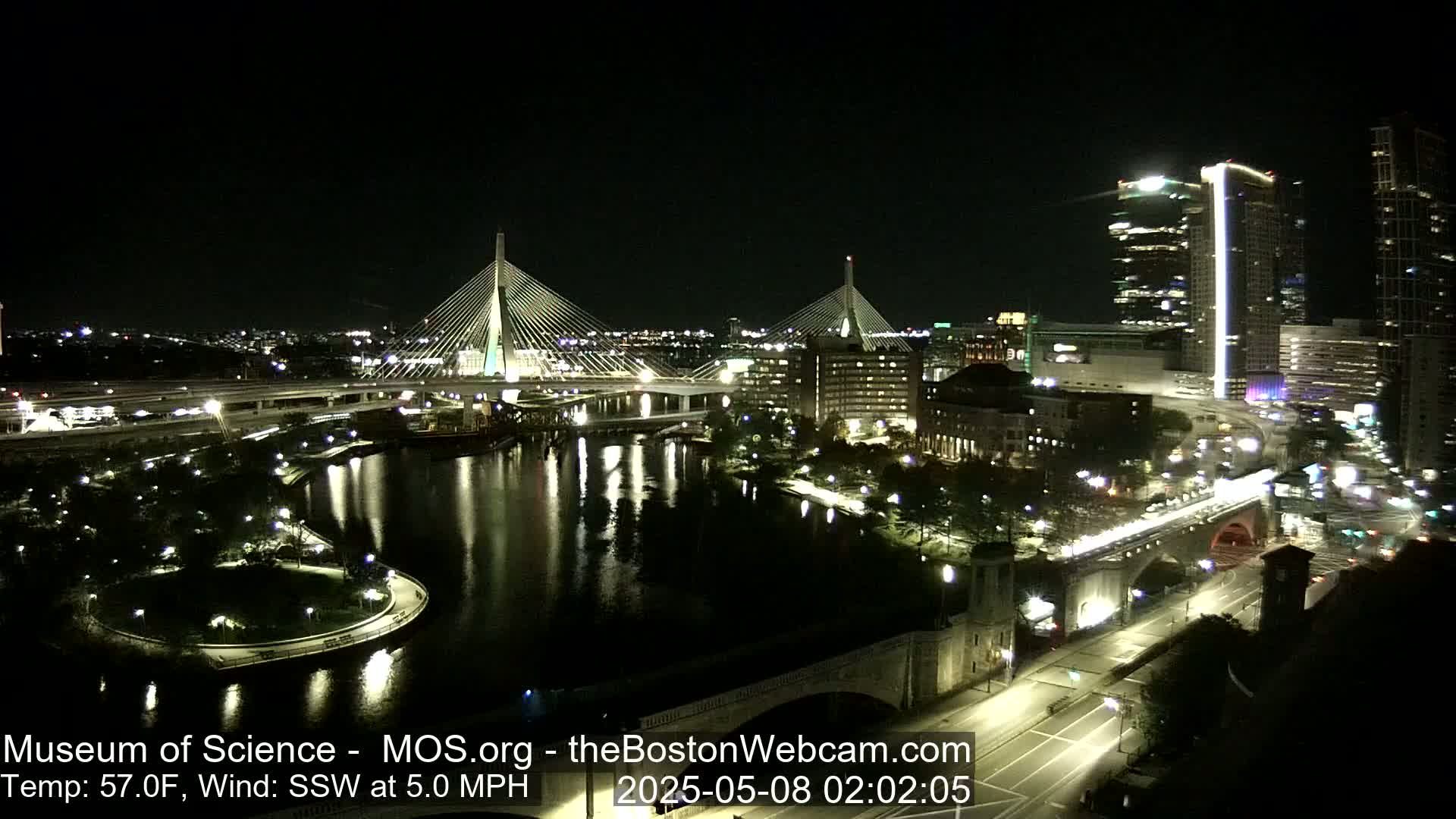 A nighttime view of two cable-stayed bridges over a dark river, with city lights reflecting in the water, under a clear sky.