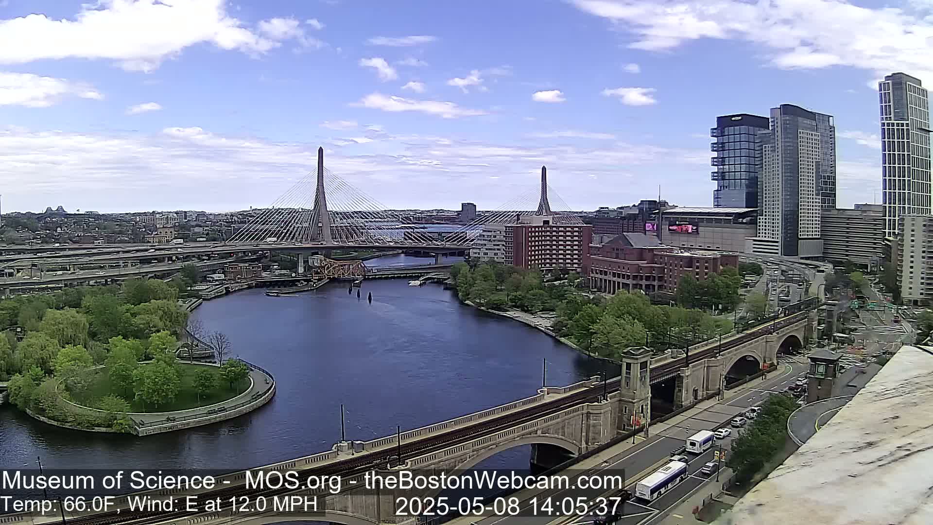 A sunny cityscape shows two cable-stayed bridges spanning a river, with a stone arch bridge in the foreground, under a partly cloudy sky.