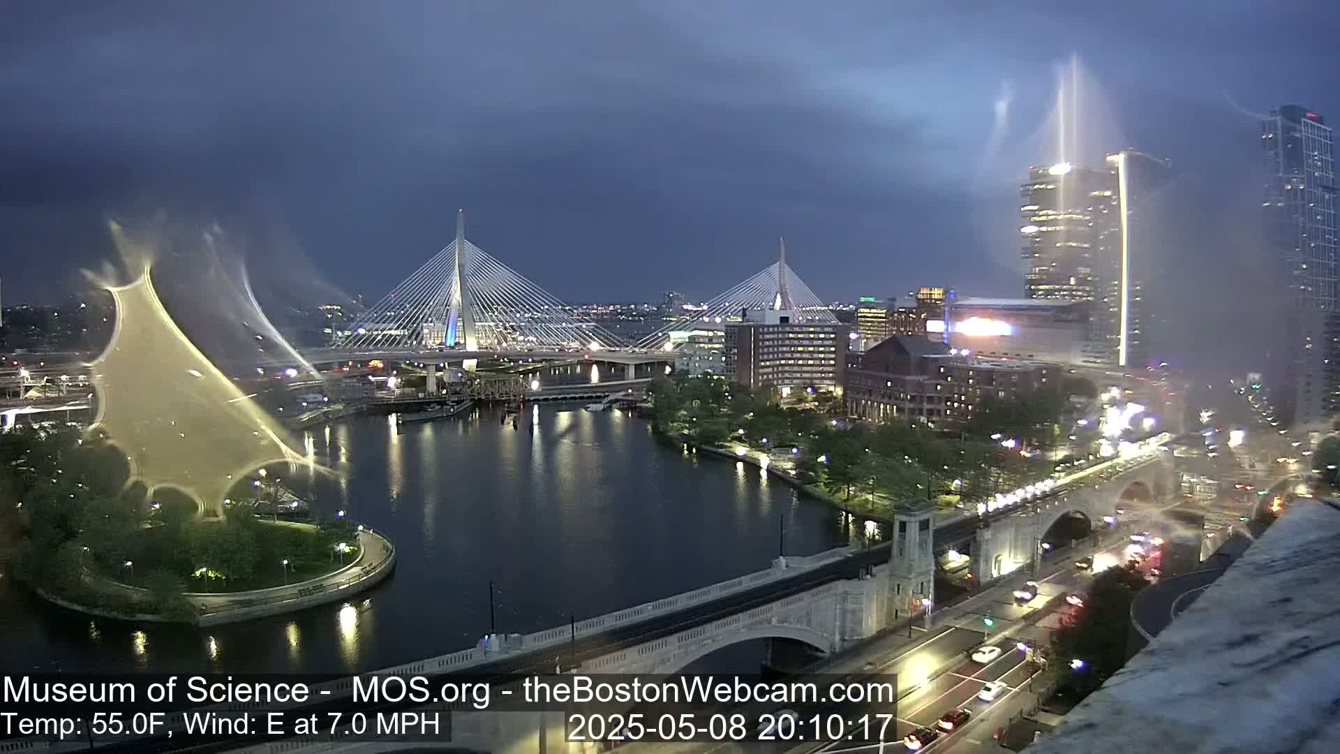 A nighttime cityscape shows two cable-stayed bridges over a dark river, with city lights, buildings, and a water fountain, under a partly cloudy, breezy sky.