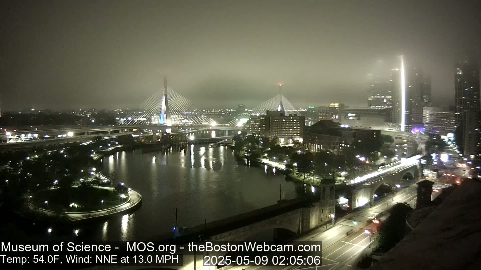 A foggy nighttime cityscape shows two cable-stayed bridges illuminated over a dark river, with city lights and roadways visible.