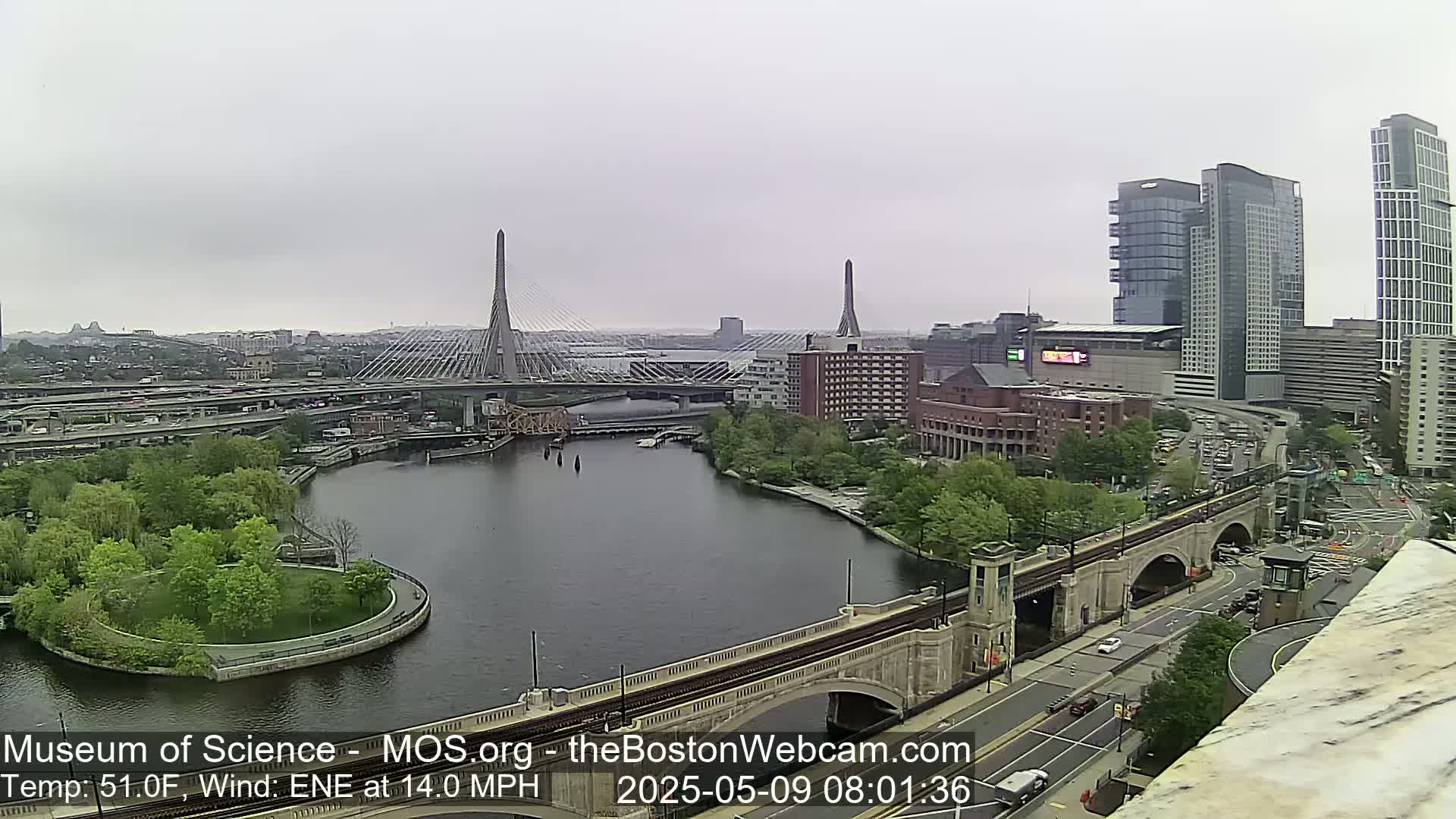 An overcast cityscape view shows a river with bridges, green spaces, and modern high-rise buildings.