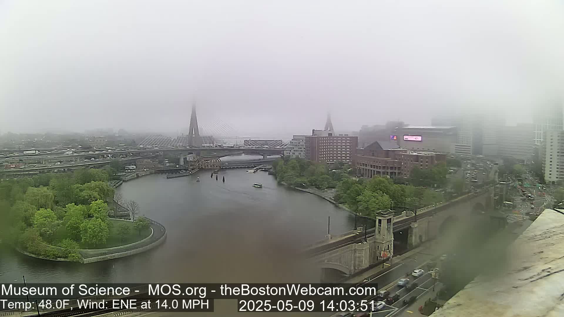 A foggy cityscape is seen from an elevated viewpoint, showing a river with a bridge and several buildings, including one large arena-like structure.