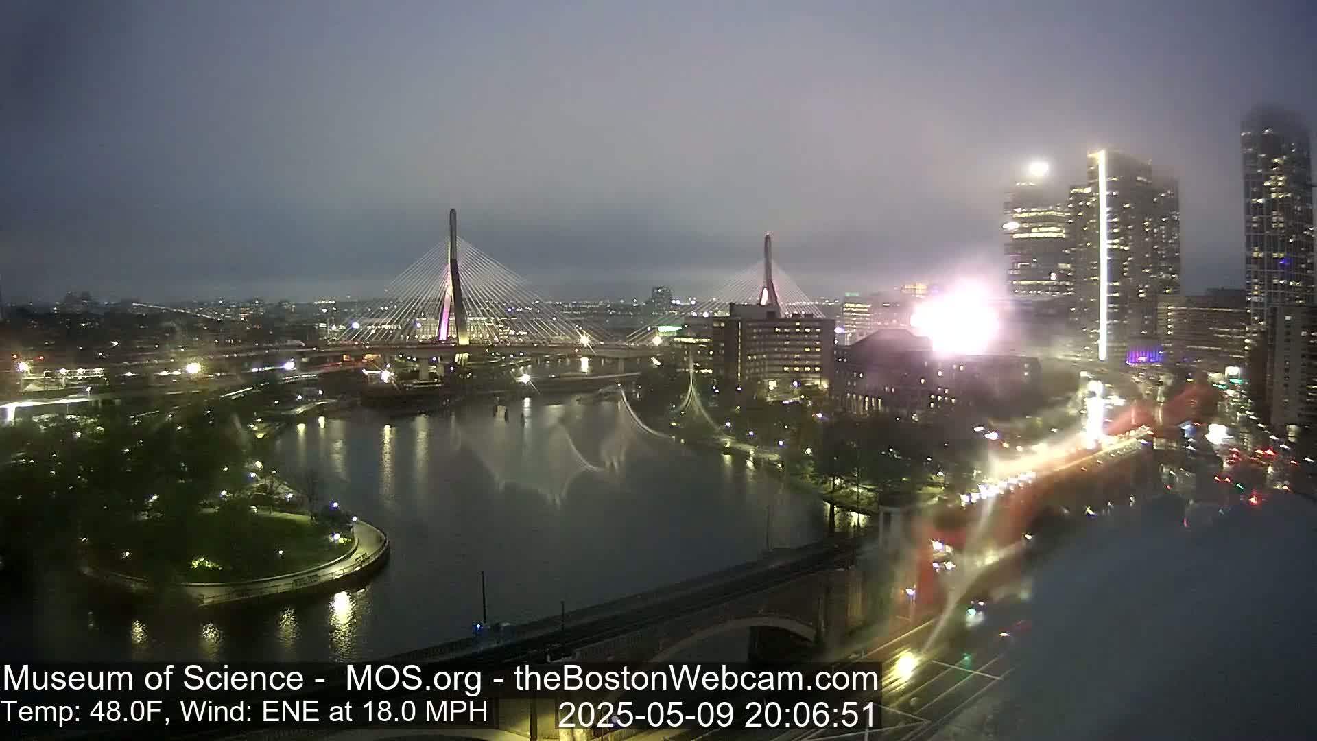 A nighttime, misty view of two cable-stayed bridges over a river, with city lights and tall buildings in the background, under an ENE wind of 18 mph.