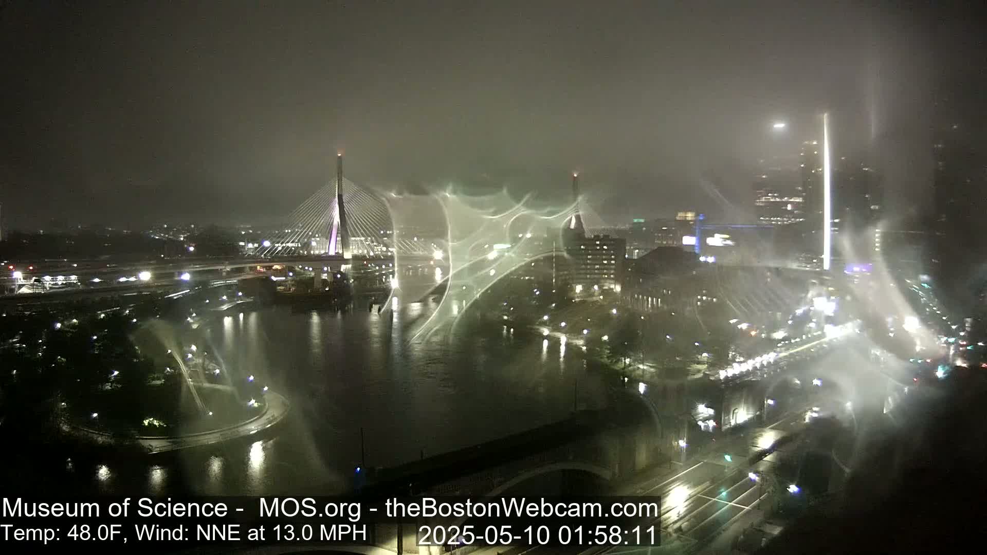 A nighttime city scene shows a bridge over a river under foggy, windy conditions with lights illuminating the area.
