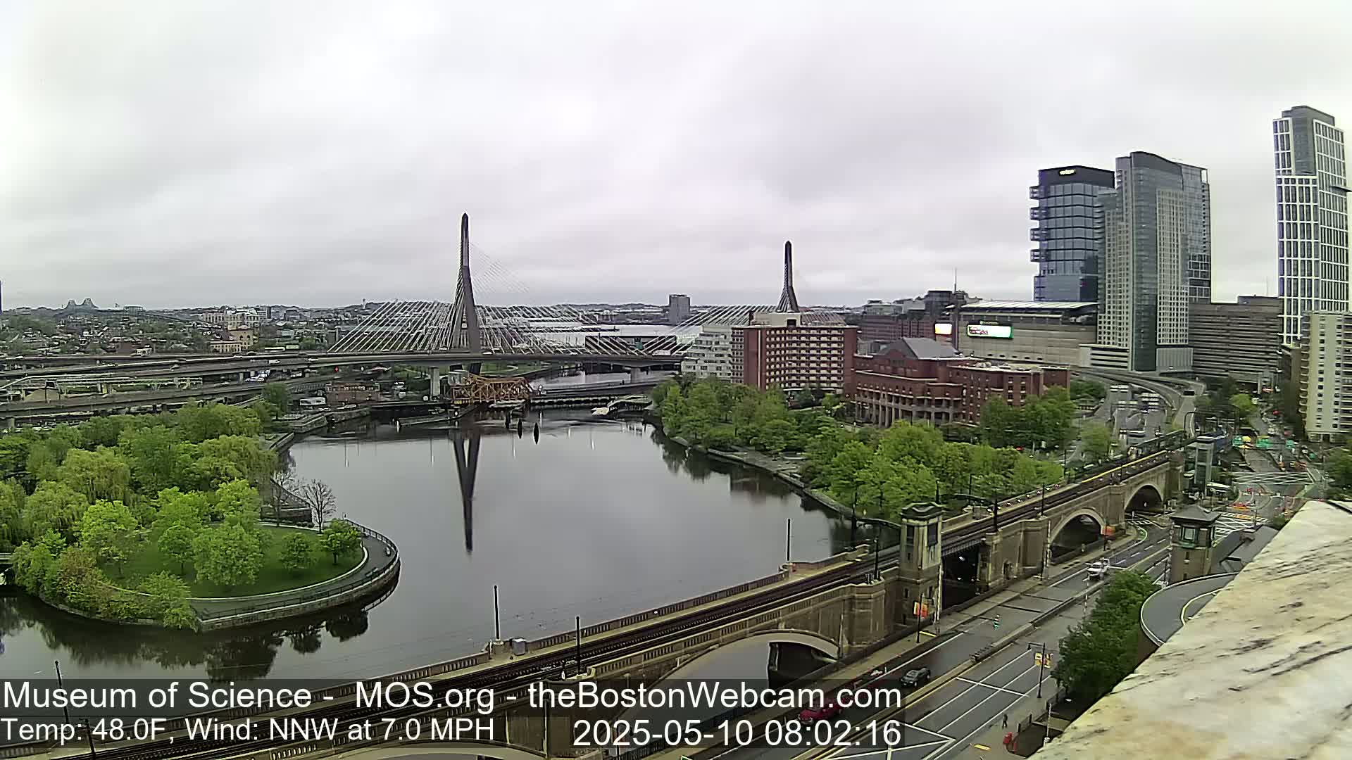 A city view on an overcast day shows two cable-stayed bridges spanning a river, with green parkland, buildings, and roadways visible.