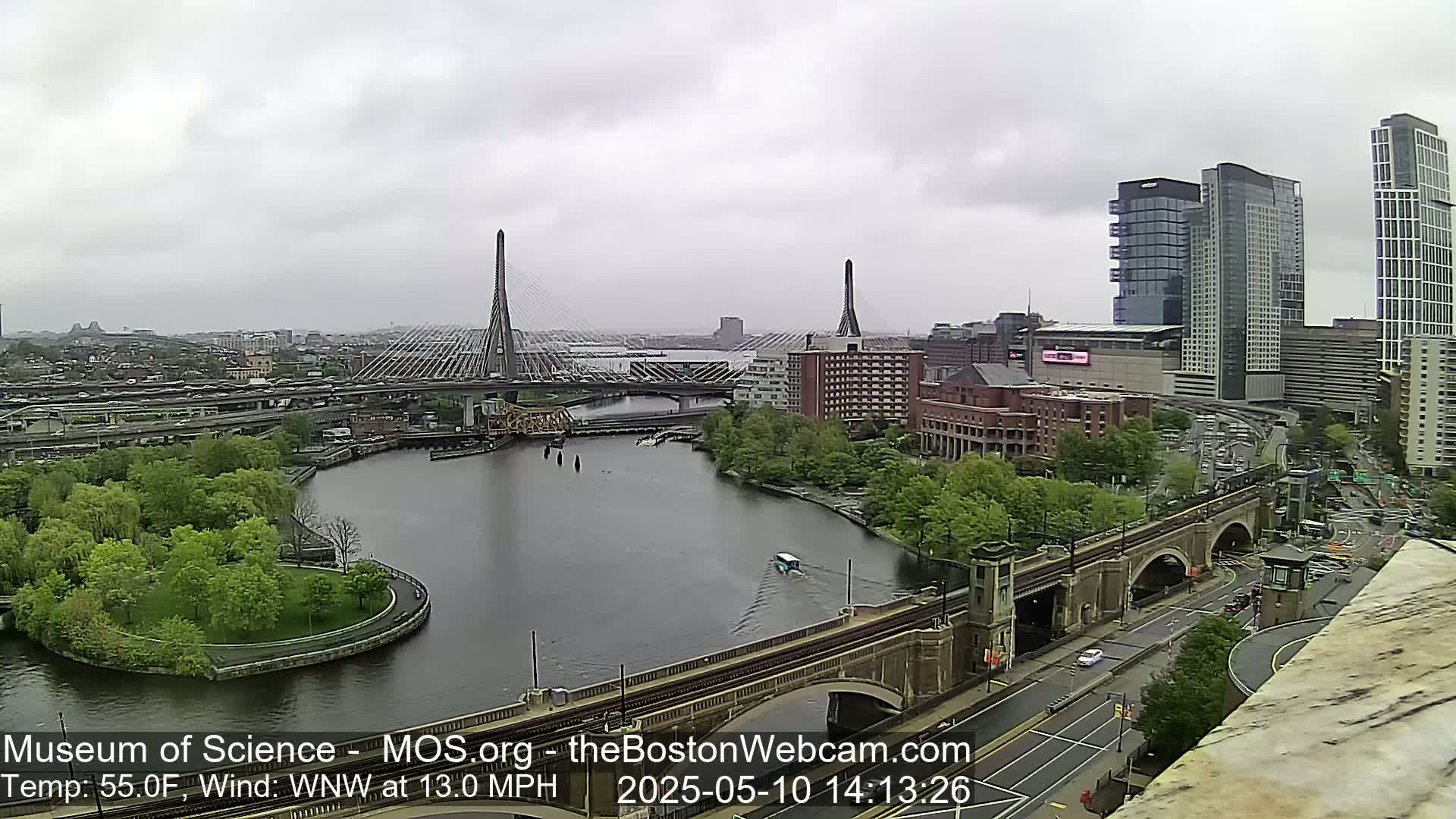 A grey, overcast day shows a cityscape with two cable-stayed bridges spanning a river, surrounded by green trees and modern high-rise buildings.
