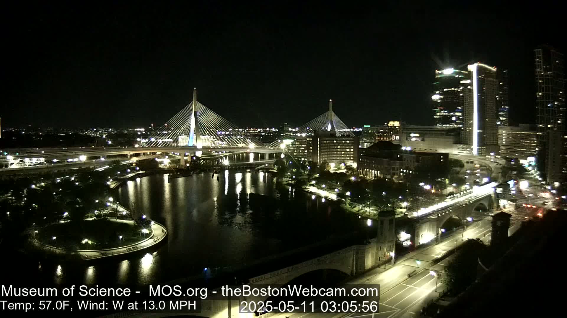 A nighttime view of two cable-stayed bridges spanning a river, with city lights and a park visible, under a clear sky with a 13 mph west wind and a temperature of 57°F.