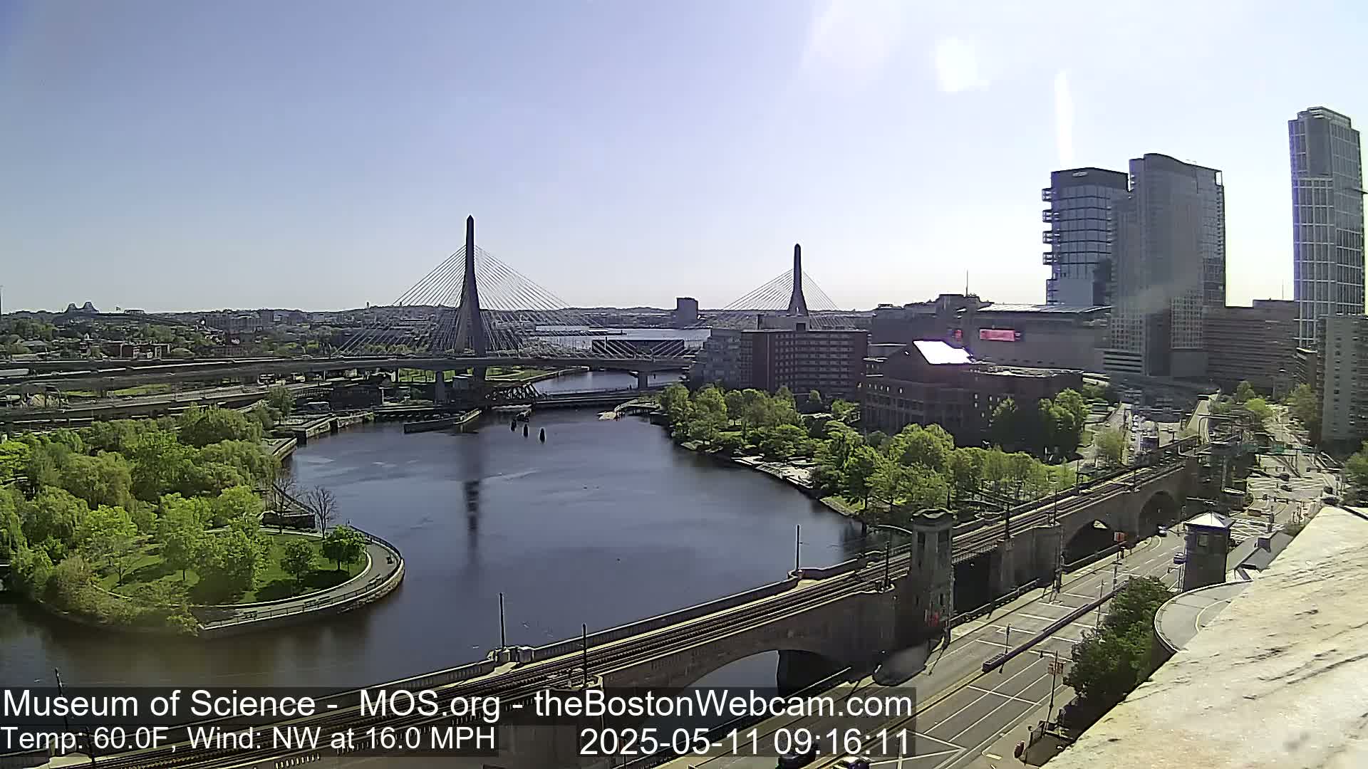 A sunny cityscape shows two cable-stayed bridges spanning a river, with green trees and modern buildings visible, under a northwest wind of 16 mph and 60°F temperature.
