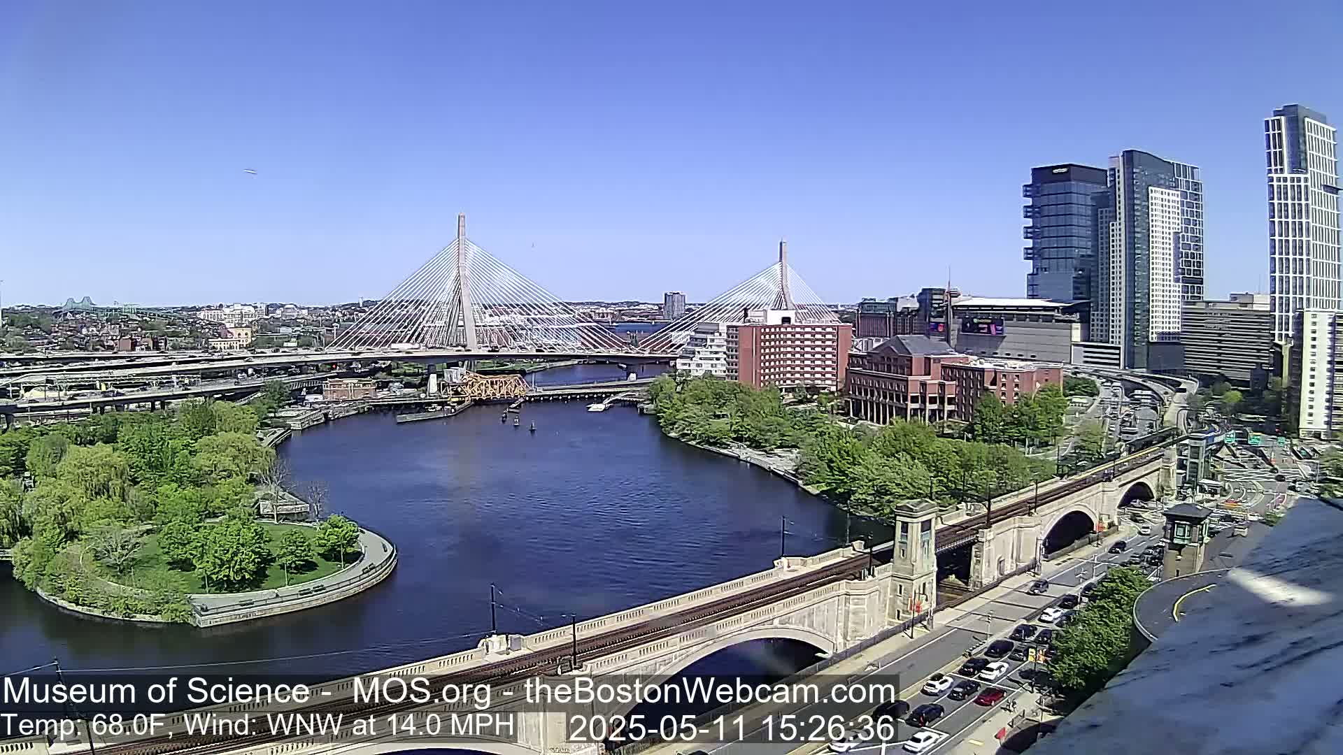 A sunny cityscape shows two cable-stayed bridges spanning a river, with modern high-rise buildings and a park visible.