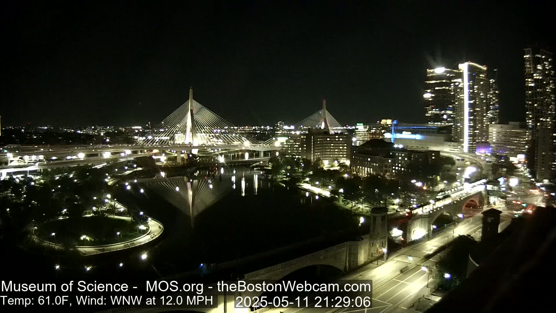 A nighttime cityscape with two illuminated cable-stayed bridges spanning a calm body of water, under a clear sky with a WNW wind of 12 mph and a temperature of 61°F.