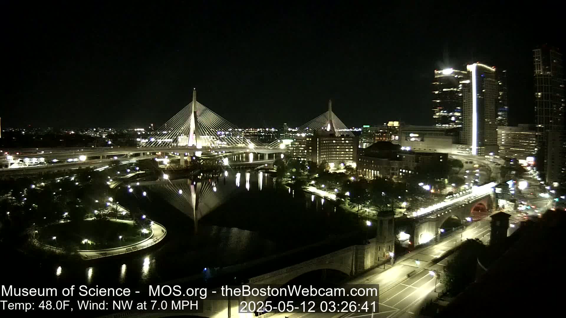 A nighttime cityscape featuring two cable-stayed bridges over a dark river, with city lights reflecting on the water under a clear sky, and a temperature of 48 degrees Fahrenheit, with a northwest wind at 7 miles per hour.
