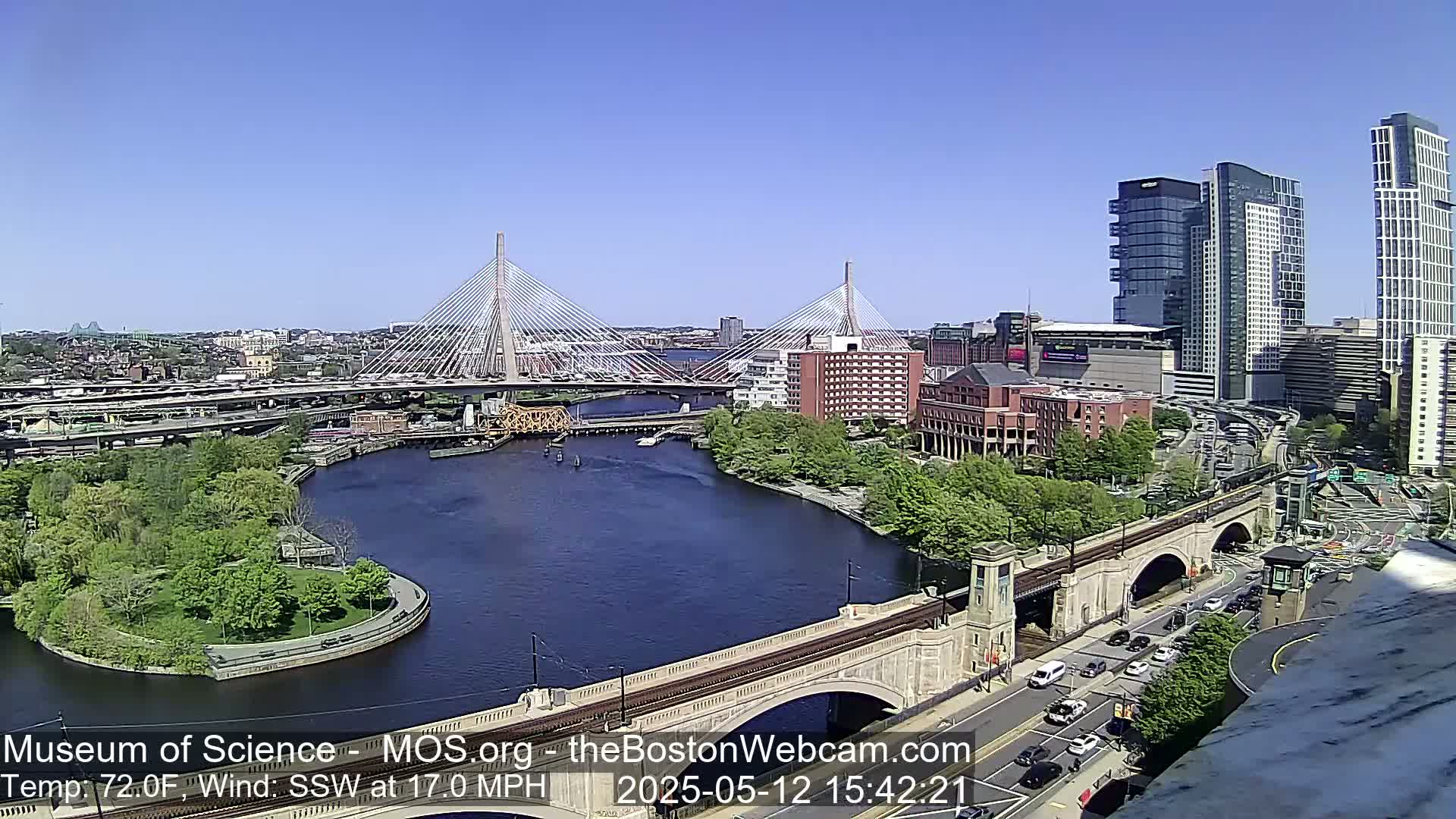 A sunny cityscape with two cable-stayed bridges spanning a river, surrounded by buildings, roadways, and green spaces.