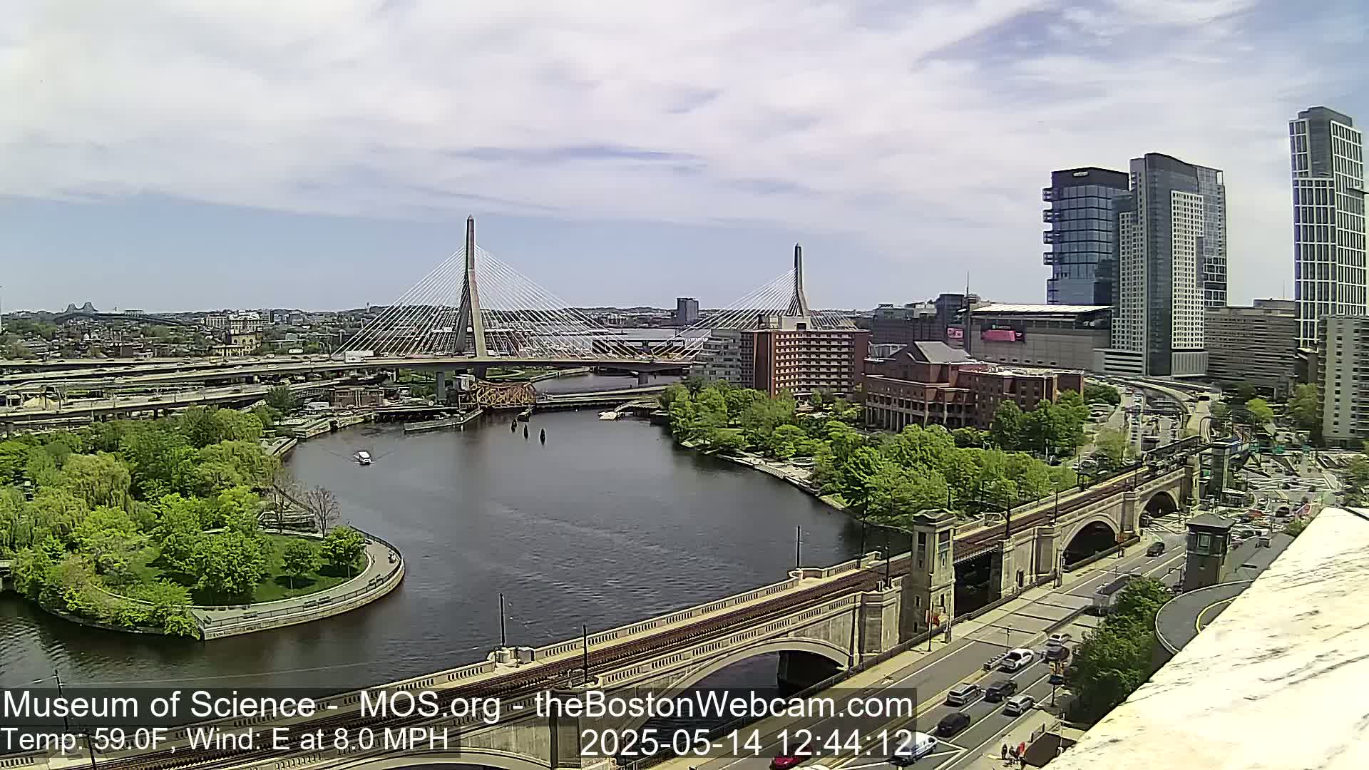 A partly cloudy day shows two cable-stayed bridges over a river, with city buildings and green spaces in the background.