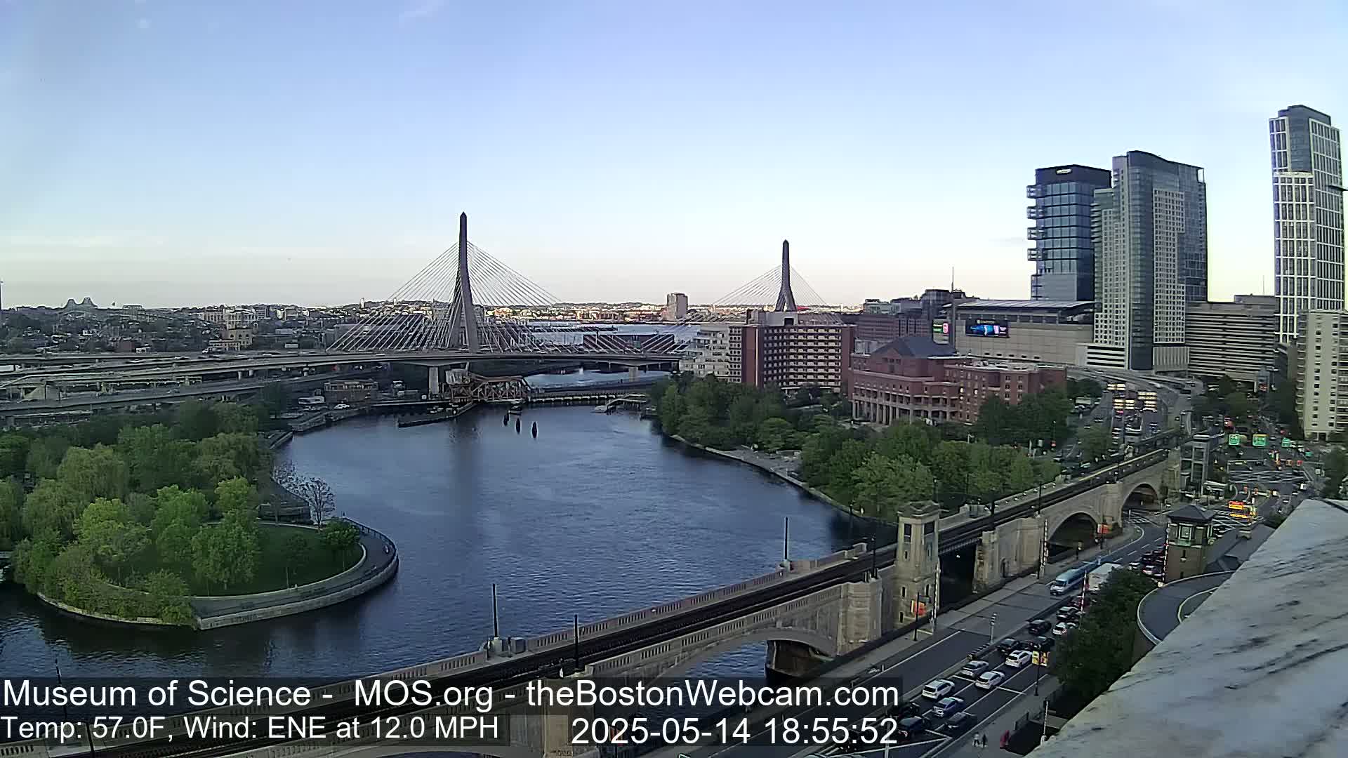 A clear evening view of a city waterfront, featuring two cable-stayed bridges, a river, and modern high-rise buildings, with a gentle ENE wind at 12 mph and a temperature of 57°F.