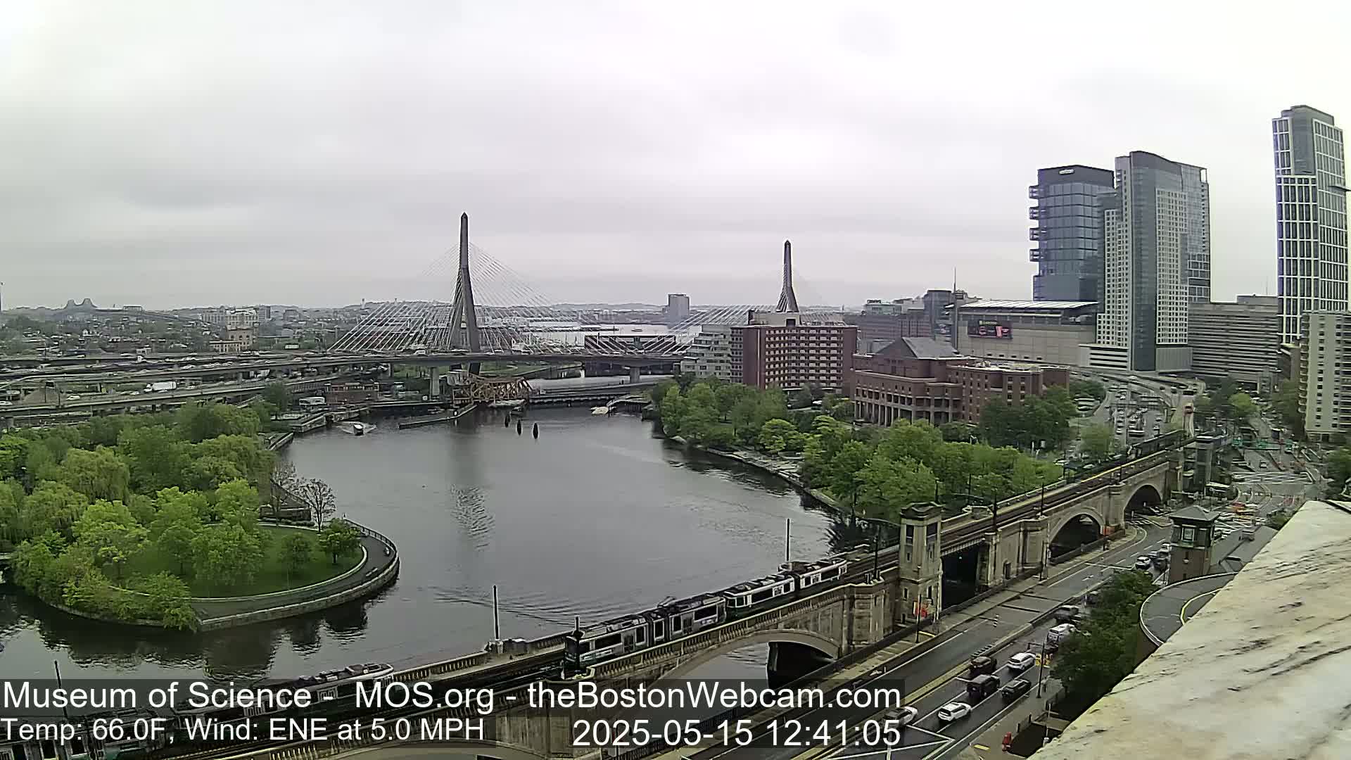 A gray day shows a city scene with two cable-stayed bridges, a river, green trees, and several tall buildings, including some high-rises.