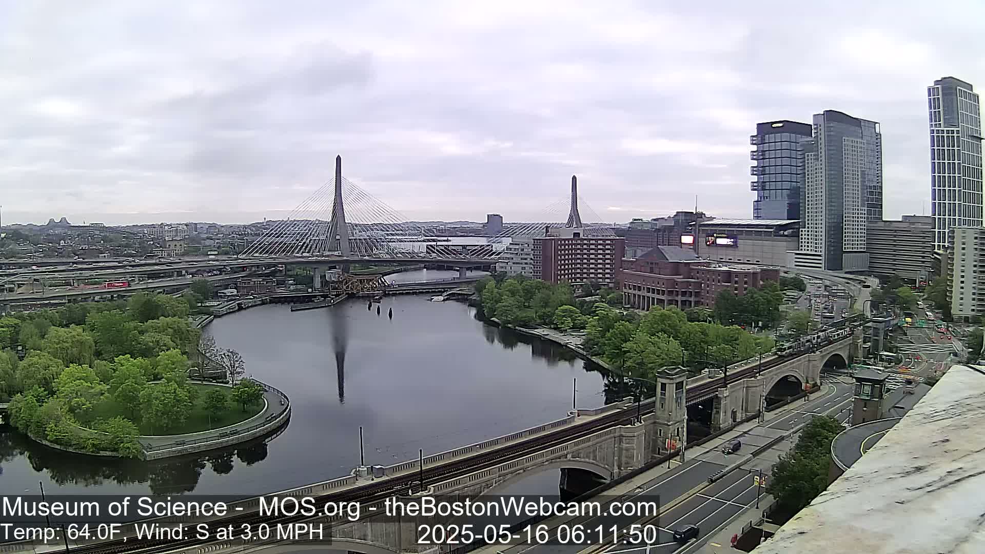 A calm, overcast day reveals a city scene with two cable-stayed bridges spanning a river, flanked by buildings and lush greenery.