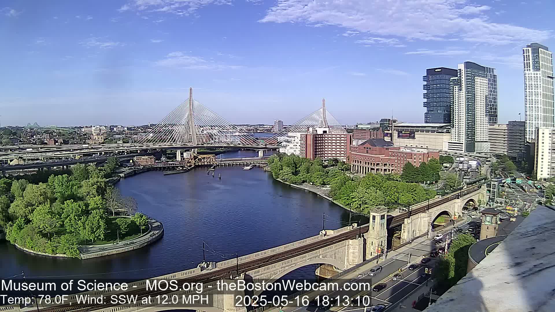 A high-angle view of a city on a sunny day with a gentle south-southwesterly wind, showing two cable-stayed bridges over a river, with several buildings and green spaces visible.