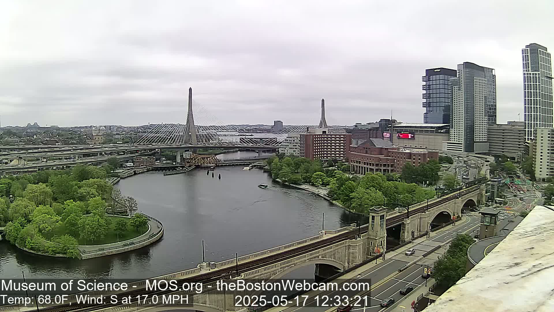 A city view on an overcast day shows two cable-stayed bridges spanning a river, with green parkland, buildings, and roadways surrounding them.