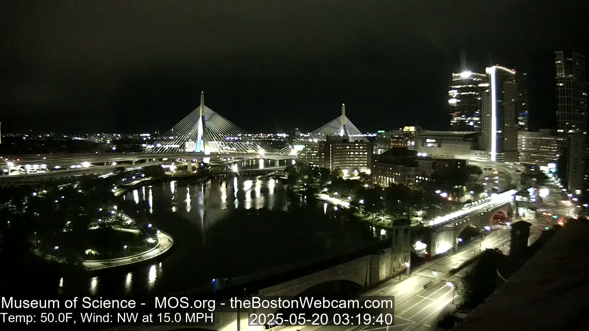 A nighttime view of two cable-stayed bridges spanning a river, with city lights and buildings illuminated under a dark sky, with a northwest wind at 15 mph and a temperature of 50°F.