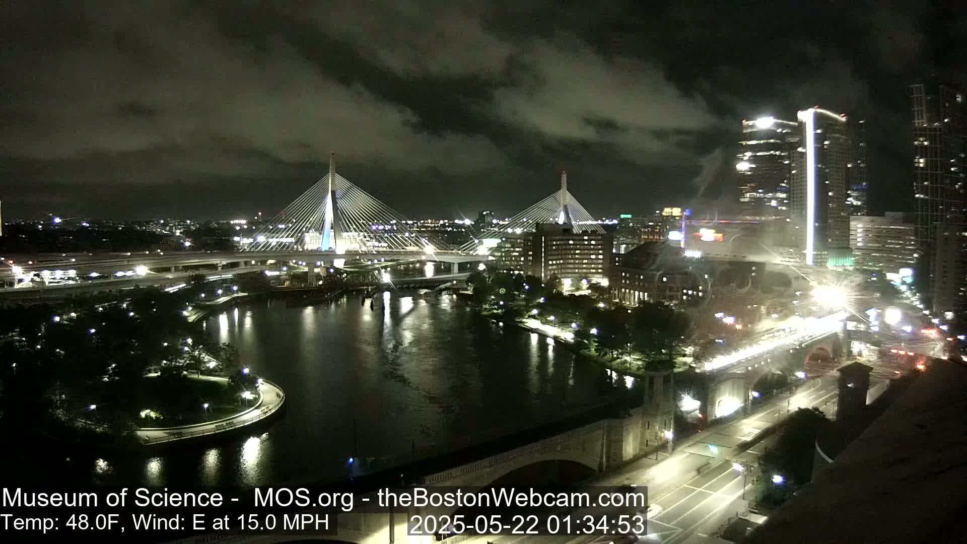 A nighttime view of two cable-stayed bridges over a dark river, with city lights and buildings visible under a cloudy sky, and a wind of 15 mph from the east.