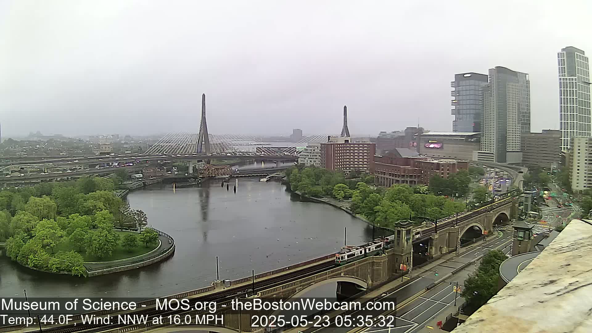 A city scene on an overcast day shows two cable-stayed bridges spanning a river, with a train passing beneath one of them.
