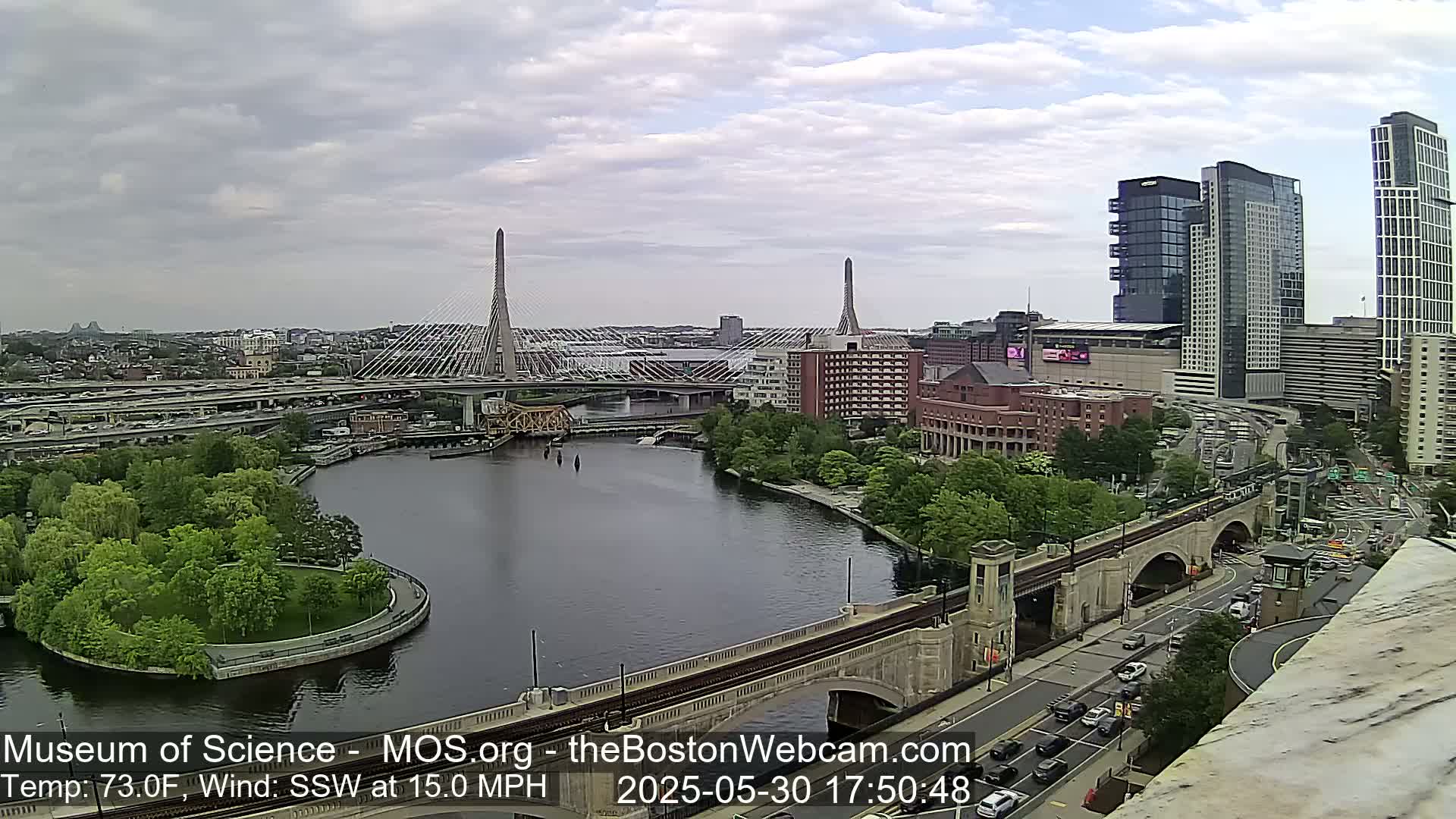A partly cloudy day shows a cityscape with two cable-stayed bridges spanning a river, surrounded by green spaces, roads, and modern high-rise buildings.