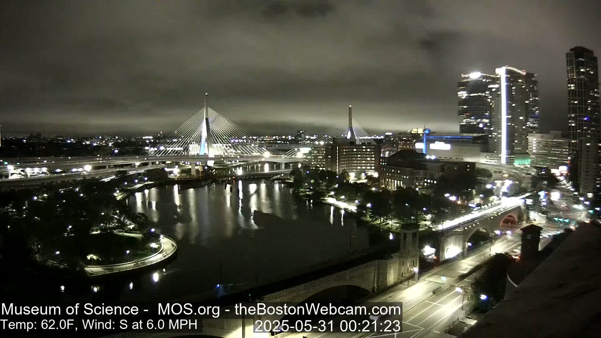 A nighttime view of a city illuminated with lights, featuring two cable-stayed bridges spanning a dark river under a cloudy sky.