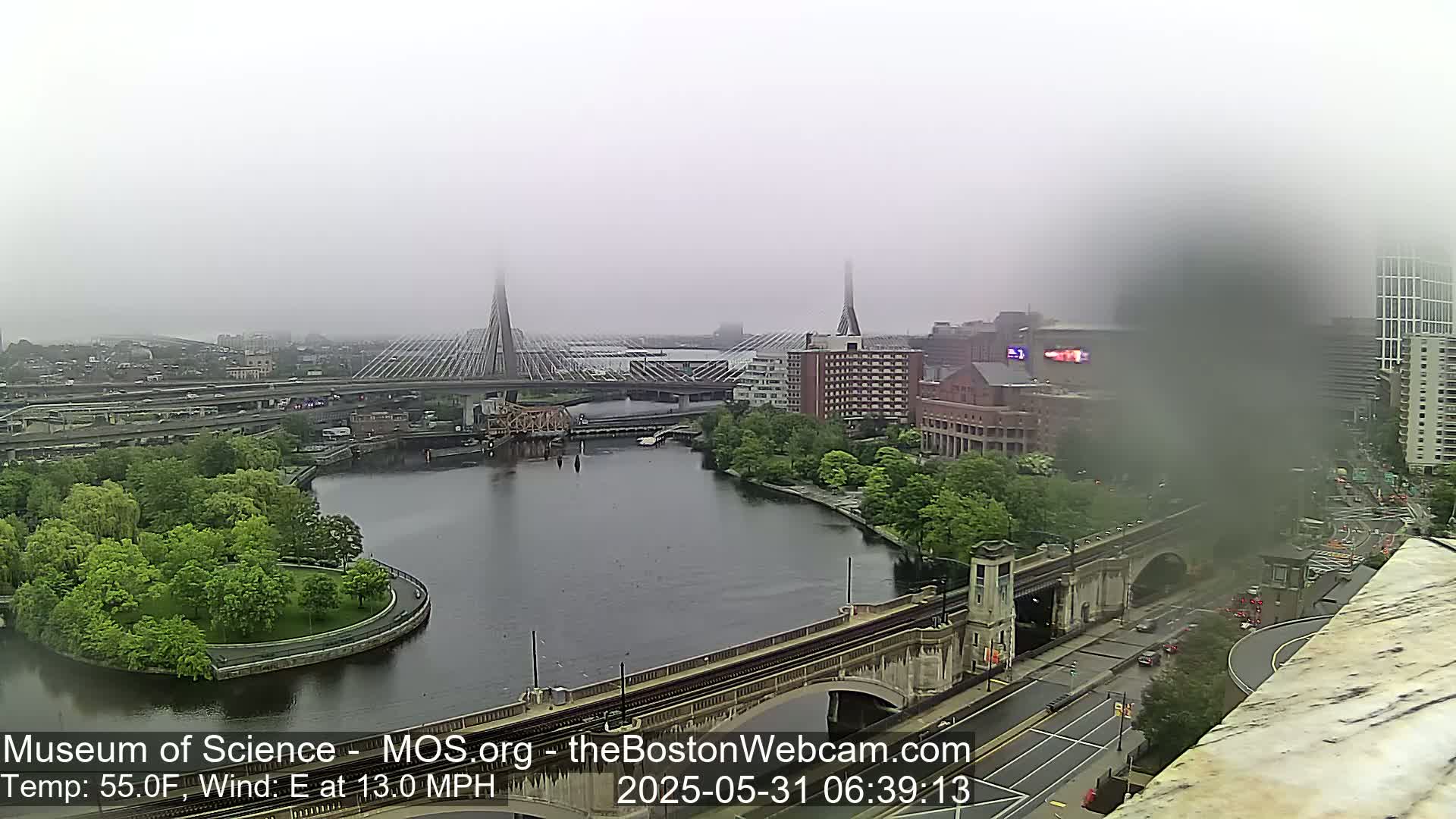 A foggy cityscape with a river, bridges, and green spaces, viewed from an elevated perspective.