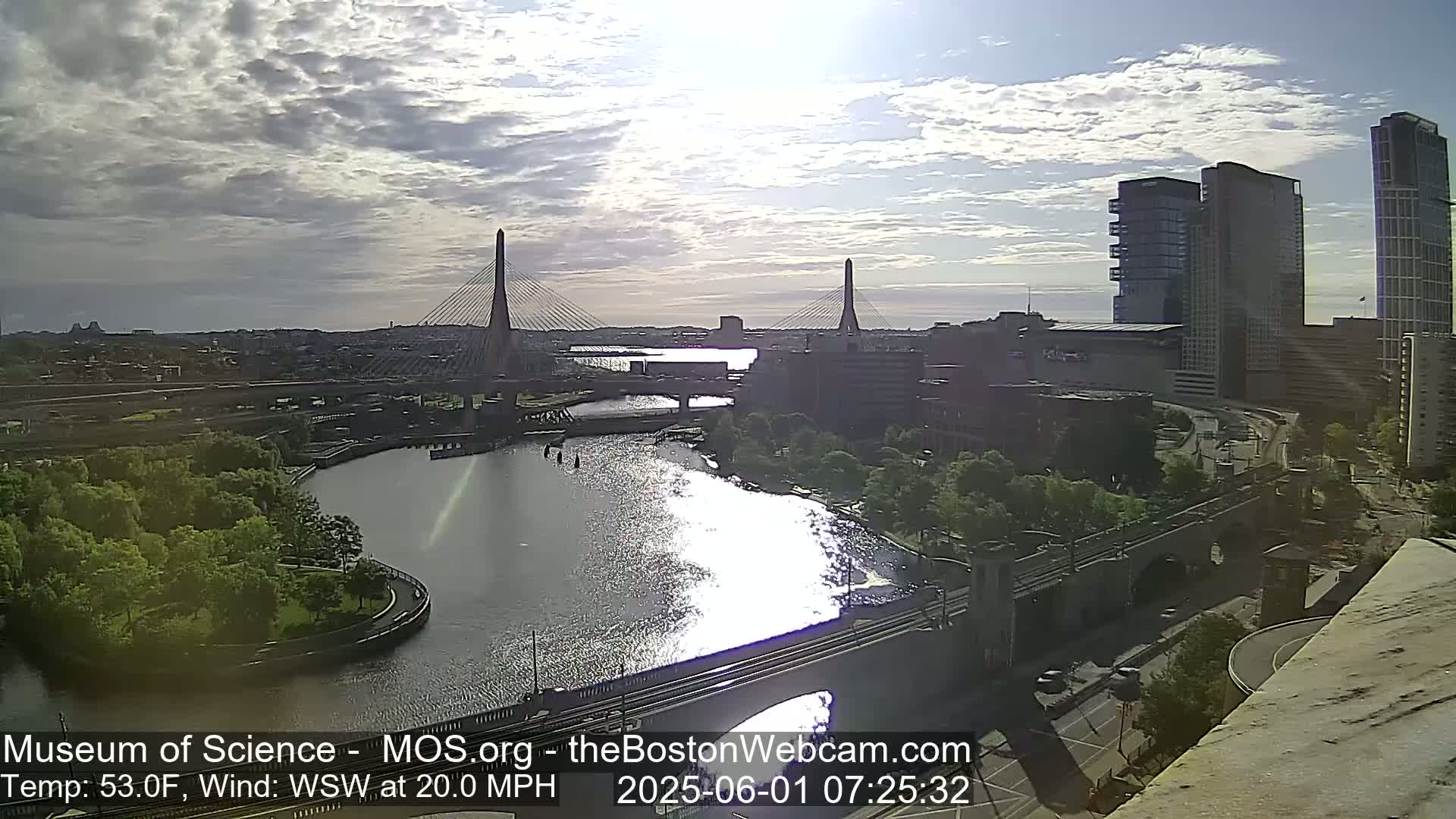 A partly cloudy sunrise scene shows two cable-stayed bridges spanning a river, with city buildings and green spaces visible, under a wind of 20 mph.