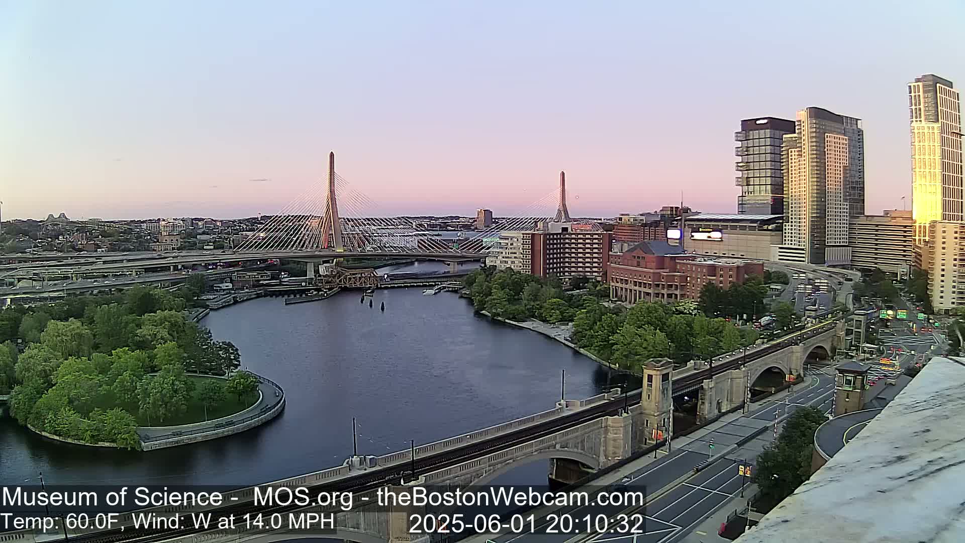 A city skyline at sunset, featuring two cable-stayed bridges spanning a river, with a light westerly wind at 14 mph and a temperature of 60°F.
