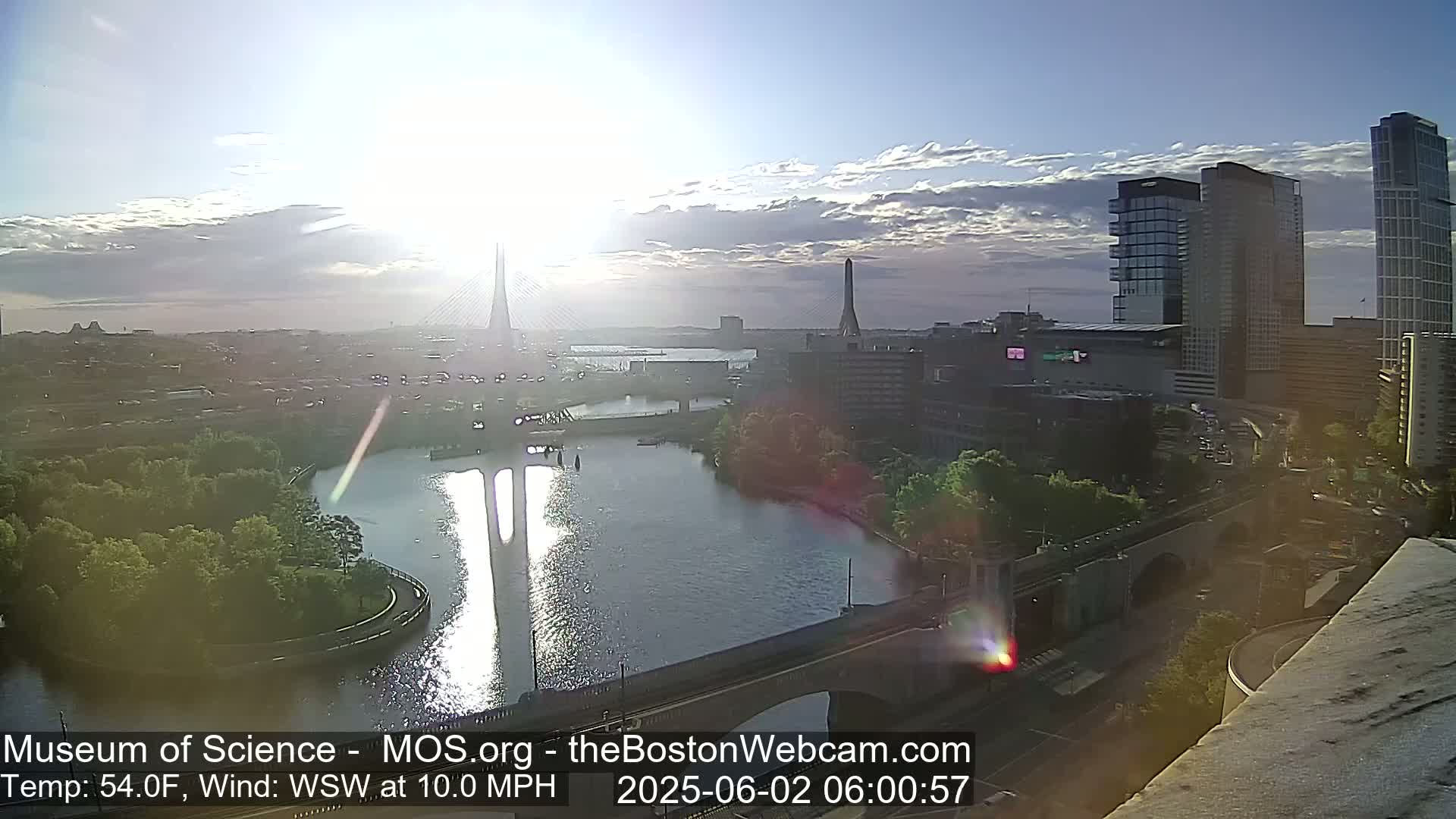 A partly cloudy sunrise illuminates a cityscape with a river, bridges, and tall buildings, with a 10 mph wind from the west-southwest.