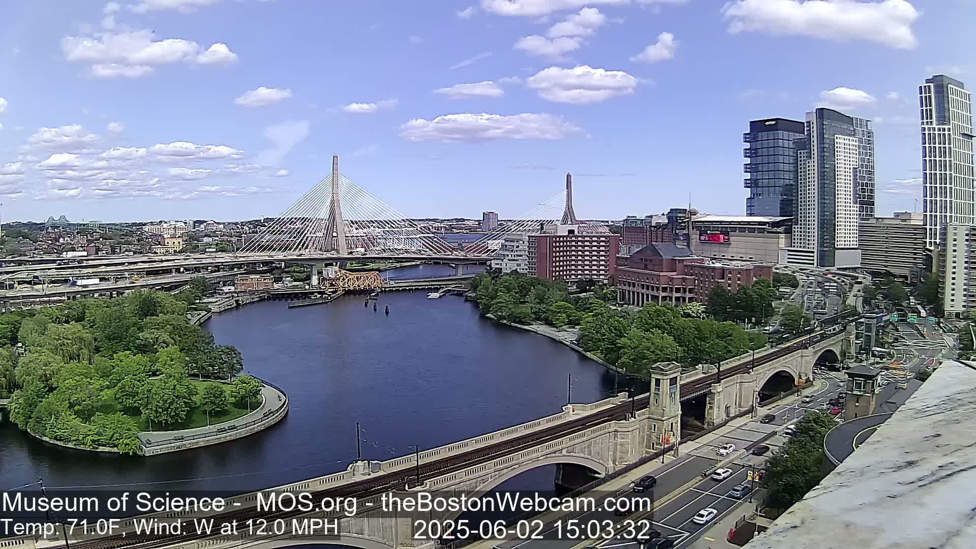 A high-angle view of a city on a partly cloudy day shows a river with two cable-stayed bridges, a stone arched bridge, and numerous buildings, including several high-rises.