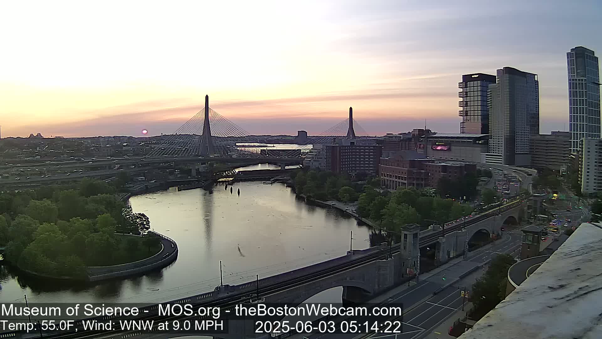 A cityscape at sunset, showing two cable-stayed bridges over a river, with calm water and a light northwest wind.