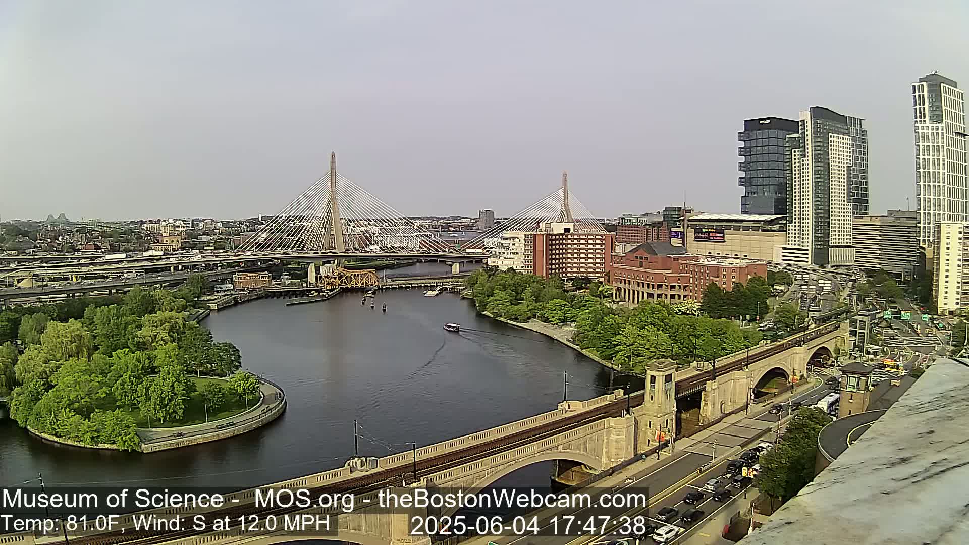 A high-angle view shows a city with two cable-stayed bridges spanning a river under a partly cloudy sky, with a south wind of 12 mph and a temperature of 81°F.