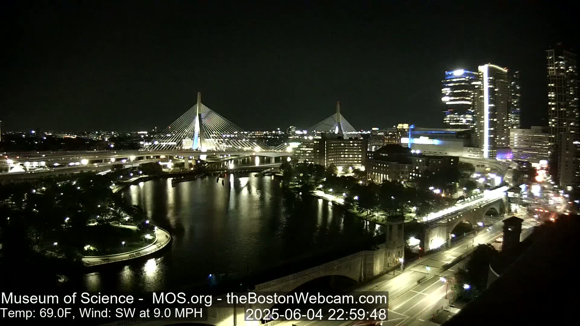 A nighttime view of a city with two illuminated cable-stayed bridges spanning a dark river, under a clear sky with a southwest wind at 9 mph and a temperature of 69°F.
