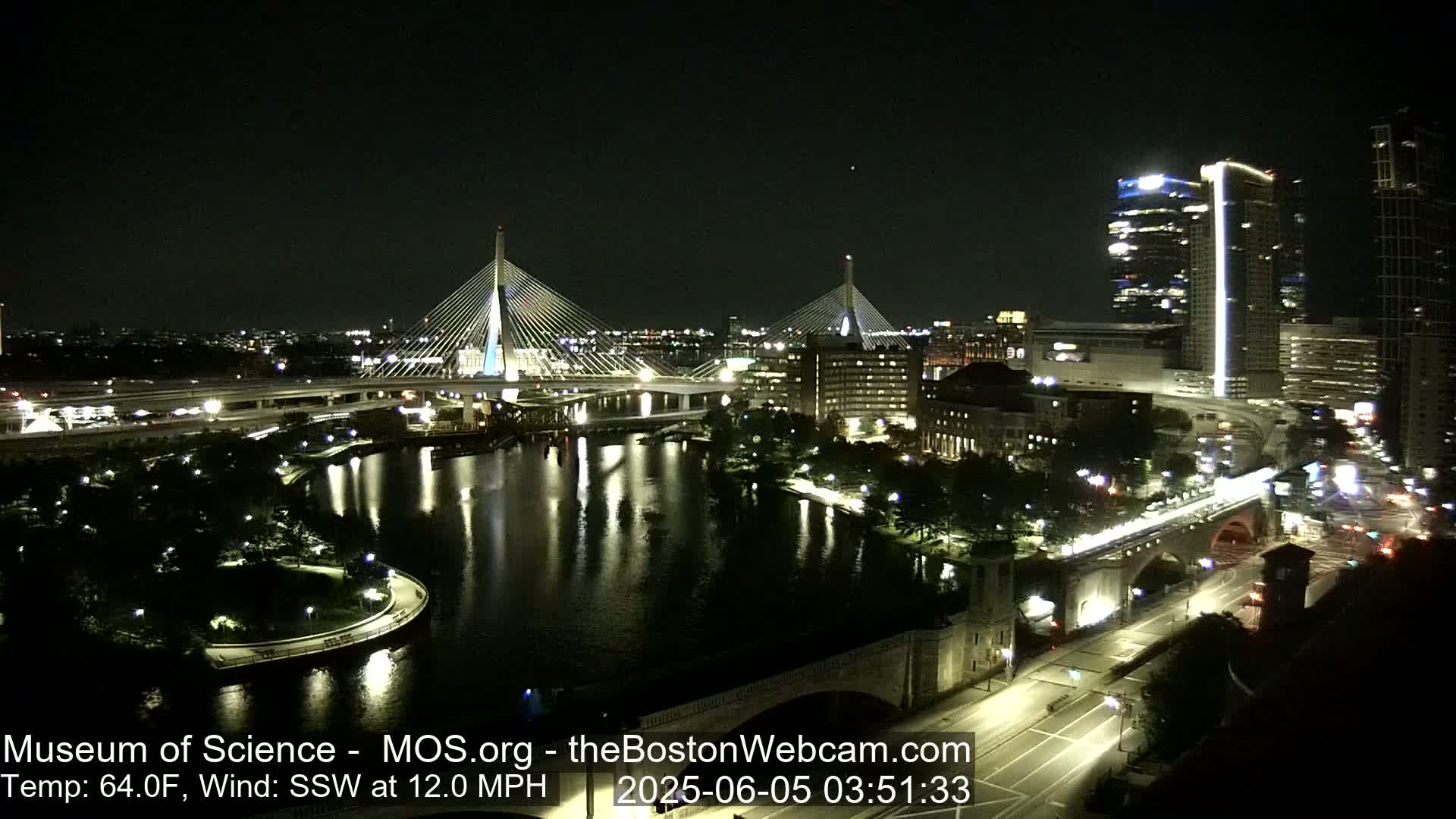 A nighttime view of a city waterfront with two cable-stayed bridges, illuminated buildings, and a calm river reflecting the city lights.
