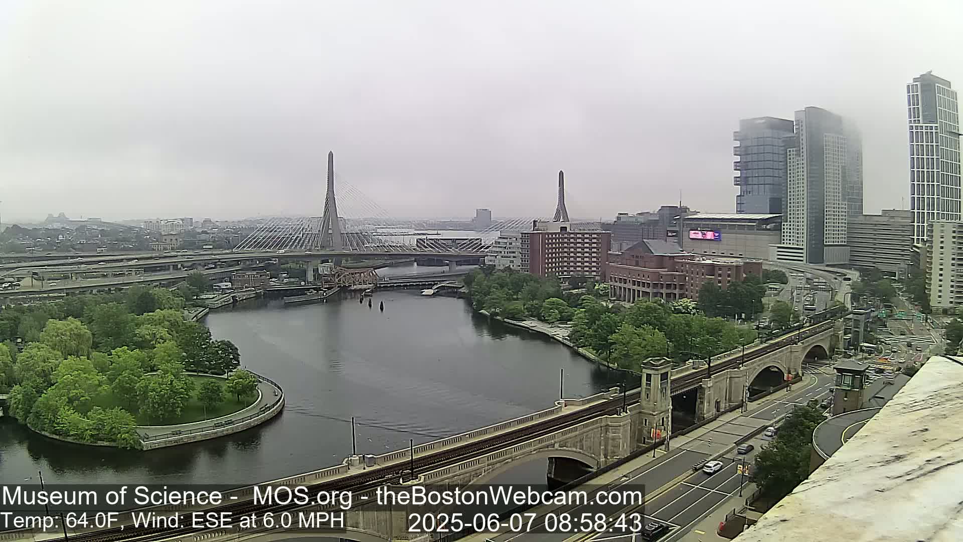 A hazy, overcast view of a city featuring two cable-stayed bridges spanning a river, with green spaces along the riverbanks and modern high-rise buildings in the background.